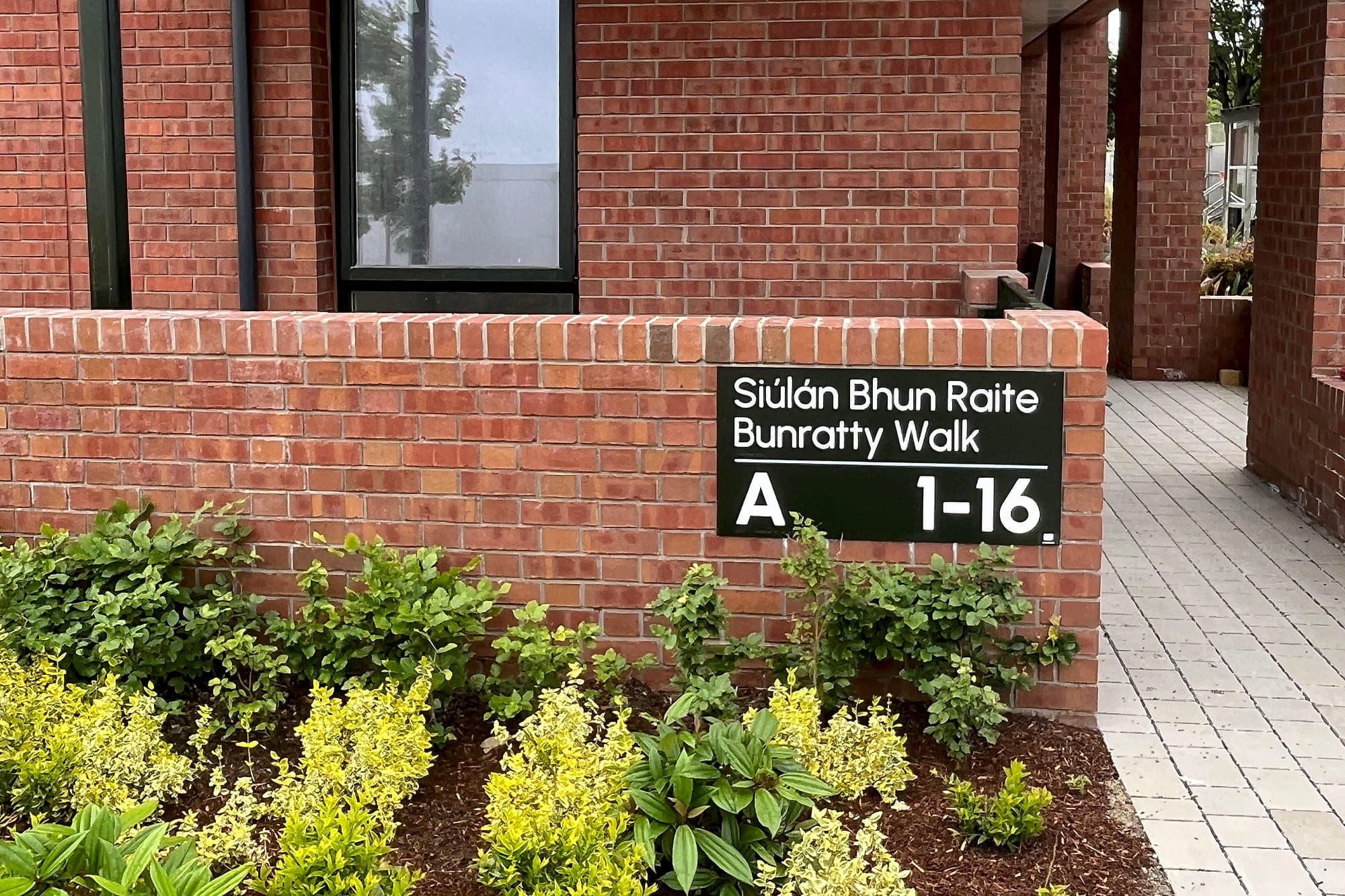 Apartment signage on a residential development: a bilingual black core sign on a brick plinth reading Siúlán Bhun Raite / Bunratty Walk over core A, apartments 1–16