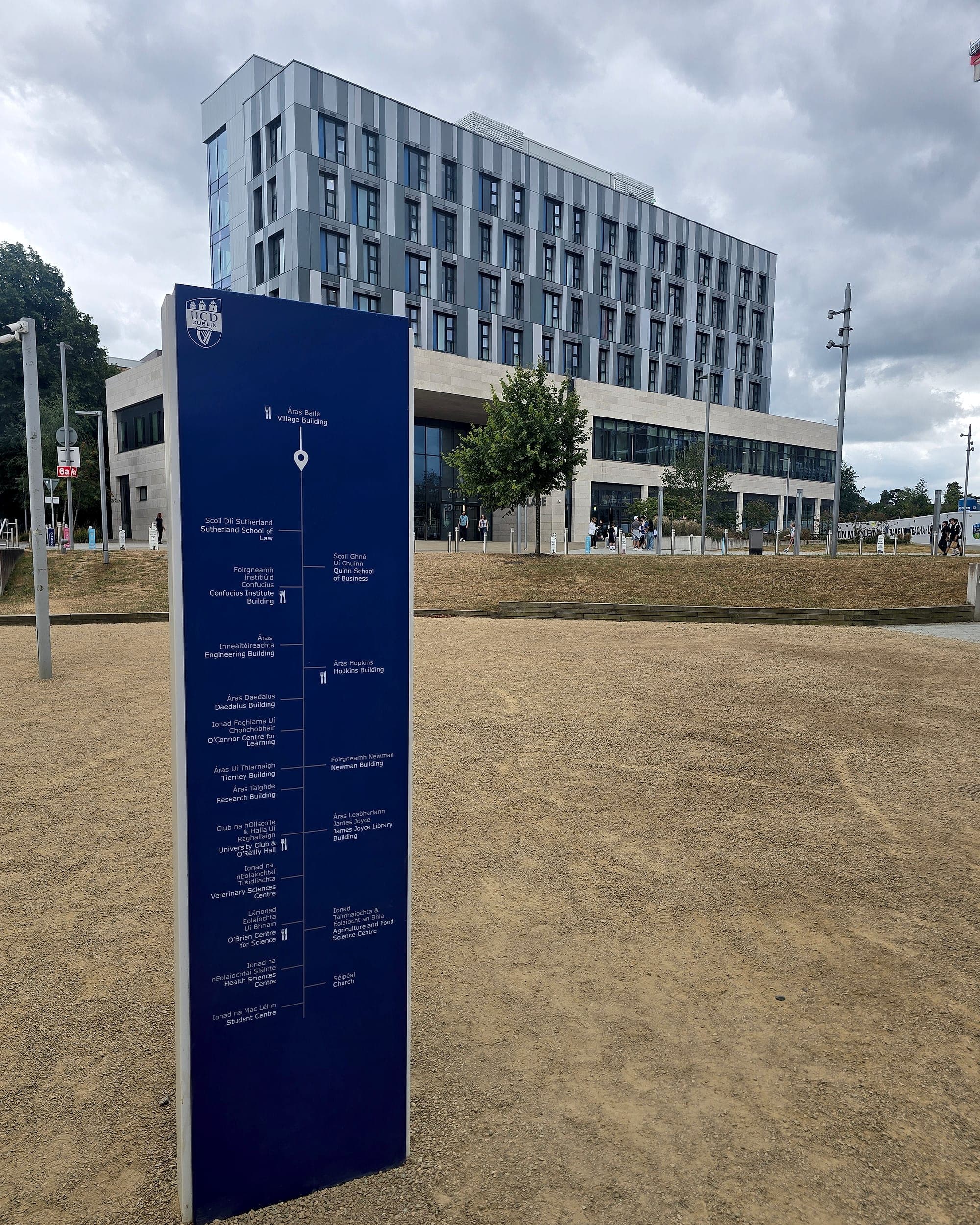 Bilingual Irish and English campus directory totem at University College Dublin, listing buildings either side of a central route line, with a modern academic block behind