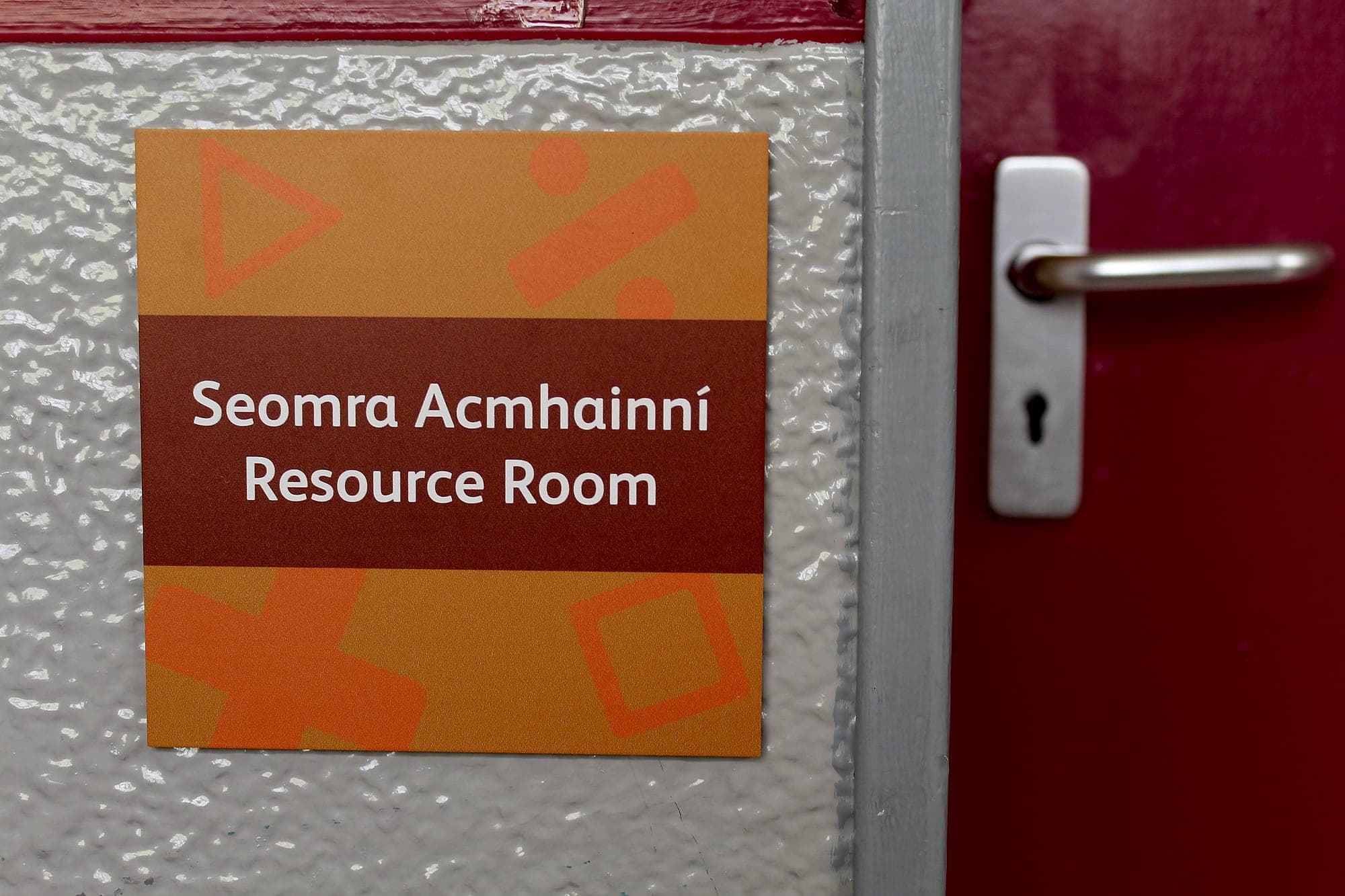 Scoil Assaim: bilingual Irish and English room identification sign reading Seomra Acmhainní / Resource Room, mounted on the wall beside a red classroom door in a school