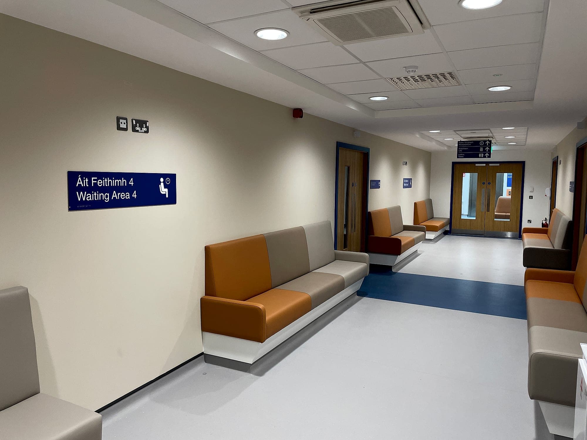 South Infirmary Victoria University Hospital: bilingual Irish and English waiting-area sign on a healthcare corridor wall, with a suspended directional sign and further wall signs visible further down the same corridor