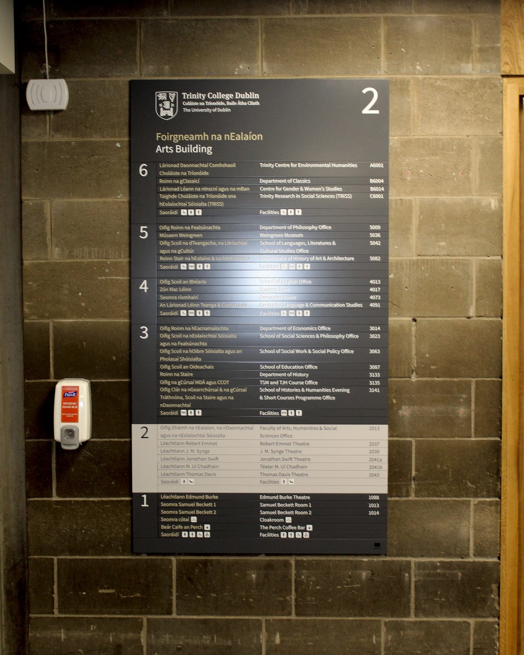 Bilingual floor directory in the Arts Building at Trinity College Dublin, listing every school, department and theatre by level in Irish and English with its room number