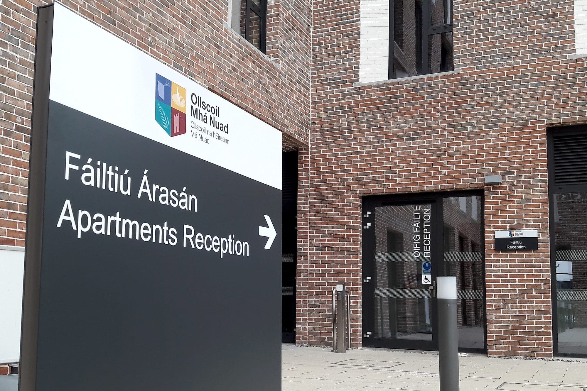 Bilingual campus directional monolith at Maynooth University pointing to apartments reception, with a matching identification plate beside the entrance door it leads to