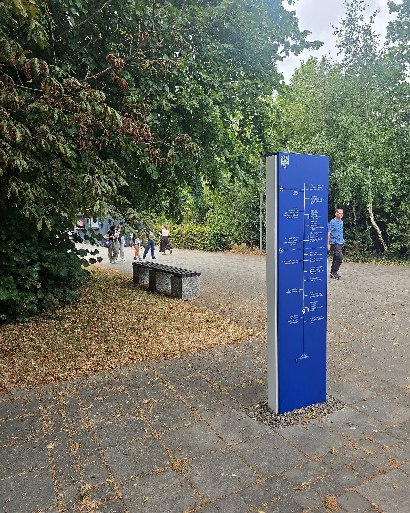 Bilingual Irish and English campus directory totem at University College Dublin, listing buildings either side of a vertical route line with a you-are-here marker, set on a gravel margin in a paved campus walkway