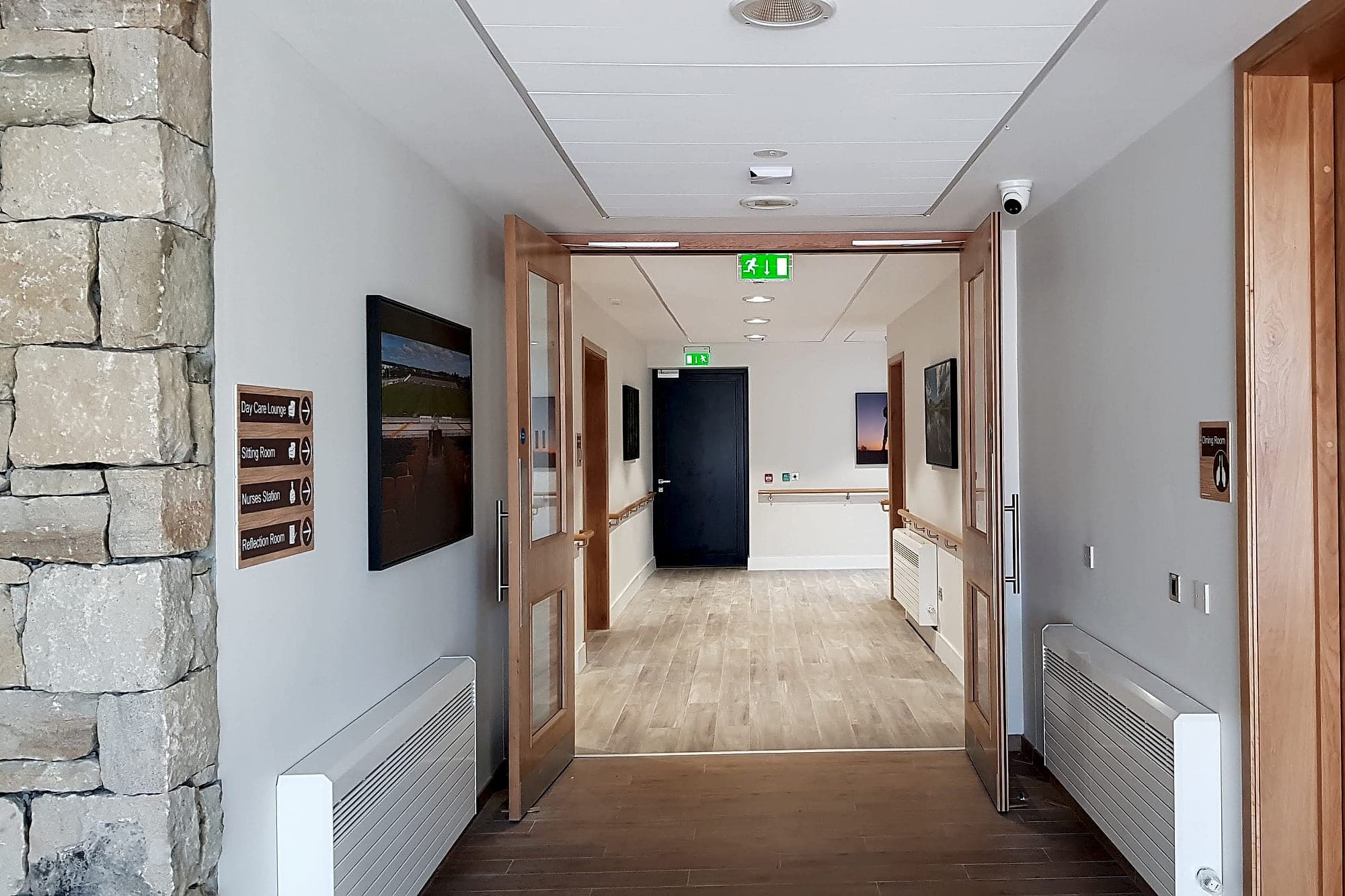 Landmarks do half the work: care home corridor with a timber-faced directional sign for day care lounge, sitting room, nurses station and reflection room, framed landscape photographs used as landmarks, handrails and a stone feature wall