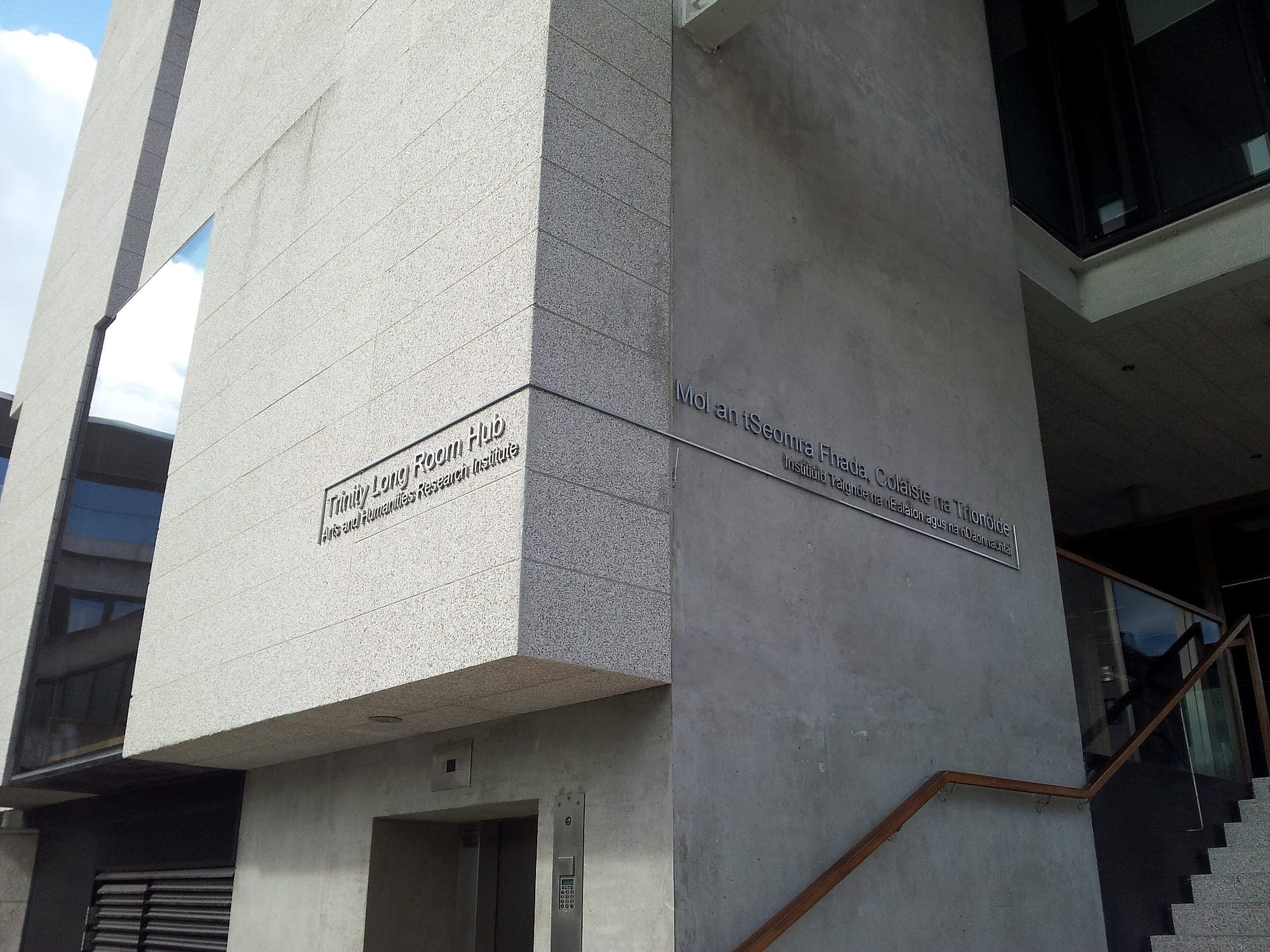 Signage installation on concrete: flat-cut stainless steel lettering pinned to a fair-faced concrete elevation and a granite return at the Trinity Long Room Hub, Trinity College Dublin