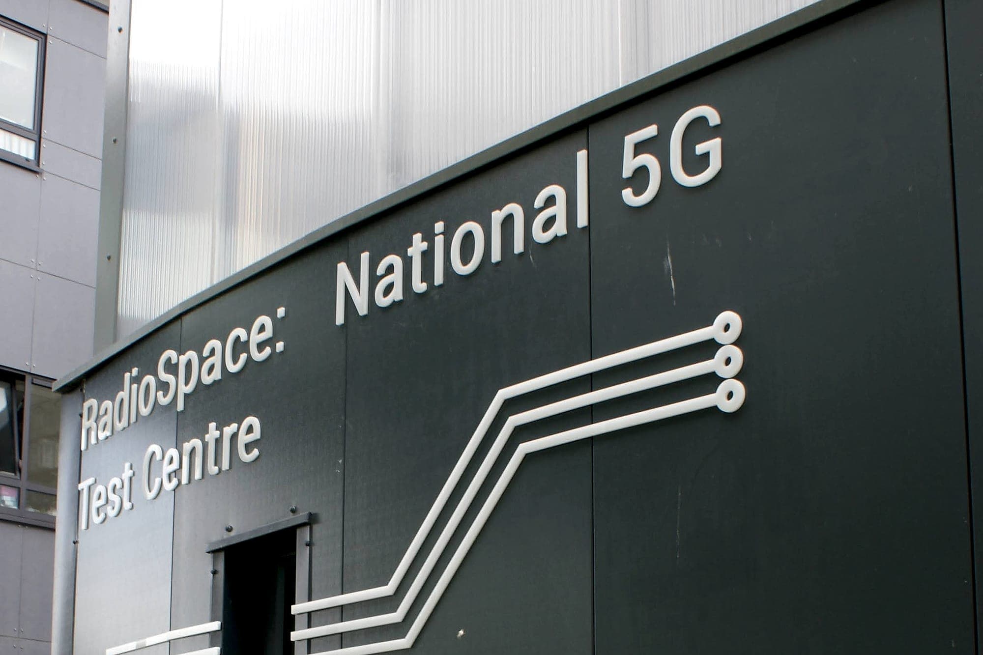 Data centre campus wayfinding: built-up building identification lettering and a circuit-trace graphic device on the metal-clad facade of a technical building
