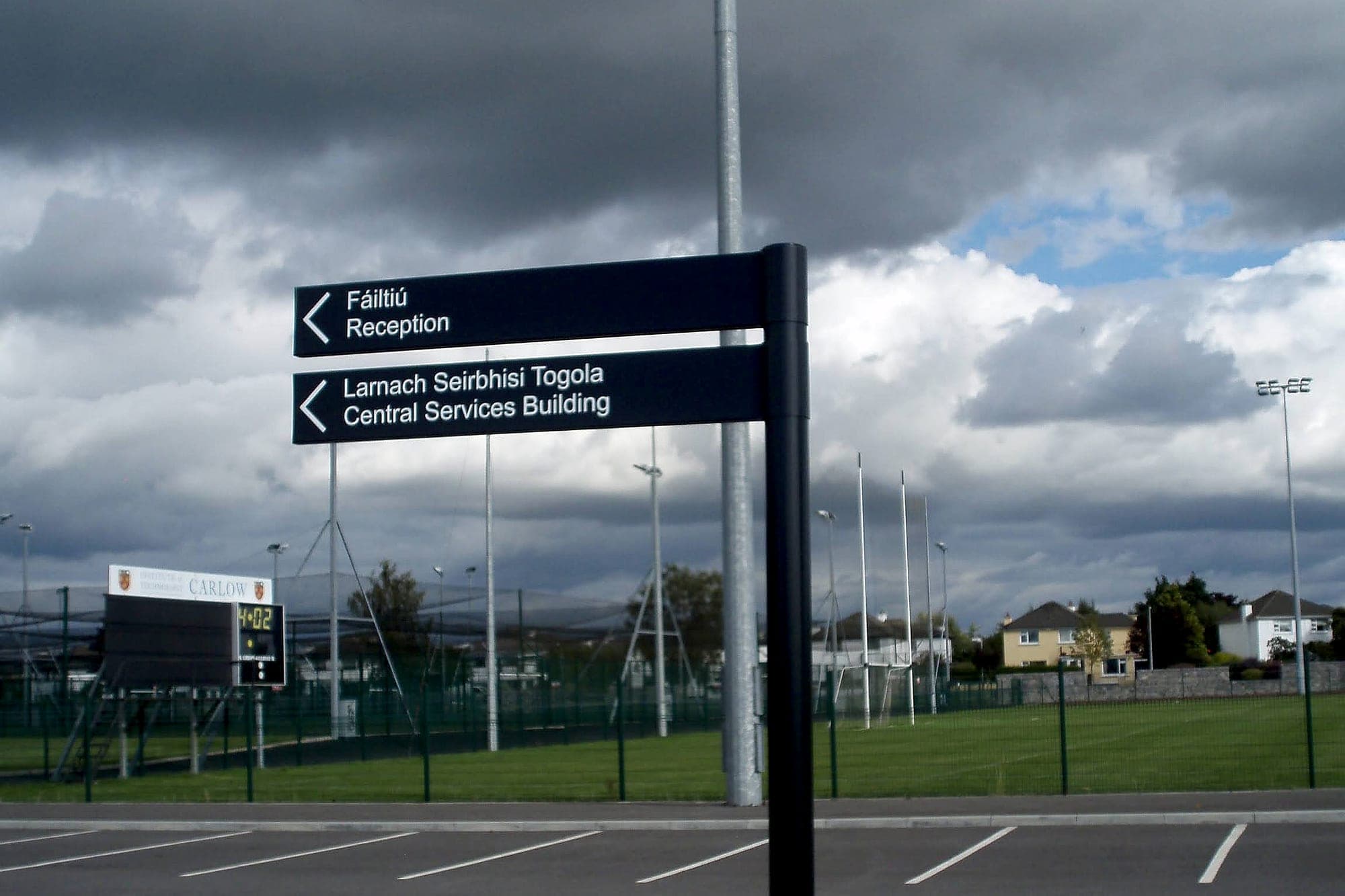 External directional signage on the approach road at IT Carlow, now SETU Carlow: a post-mounted blade sign with arrows to Reception and to the Central Services Building, seen across an empty car park