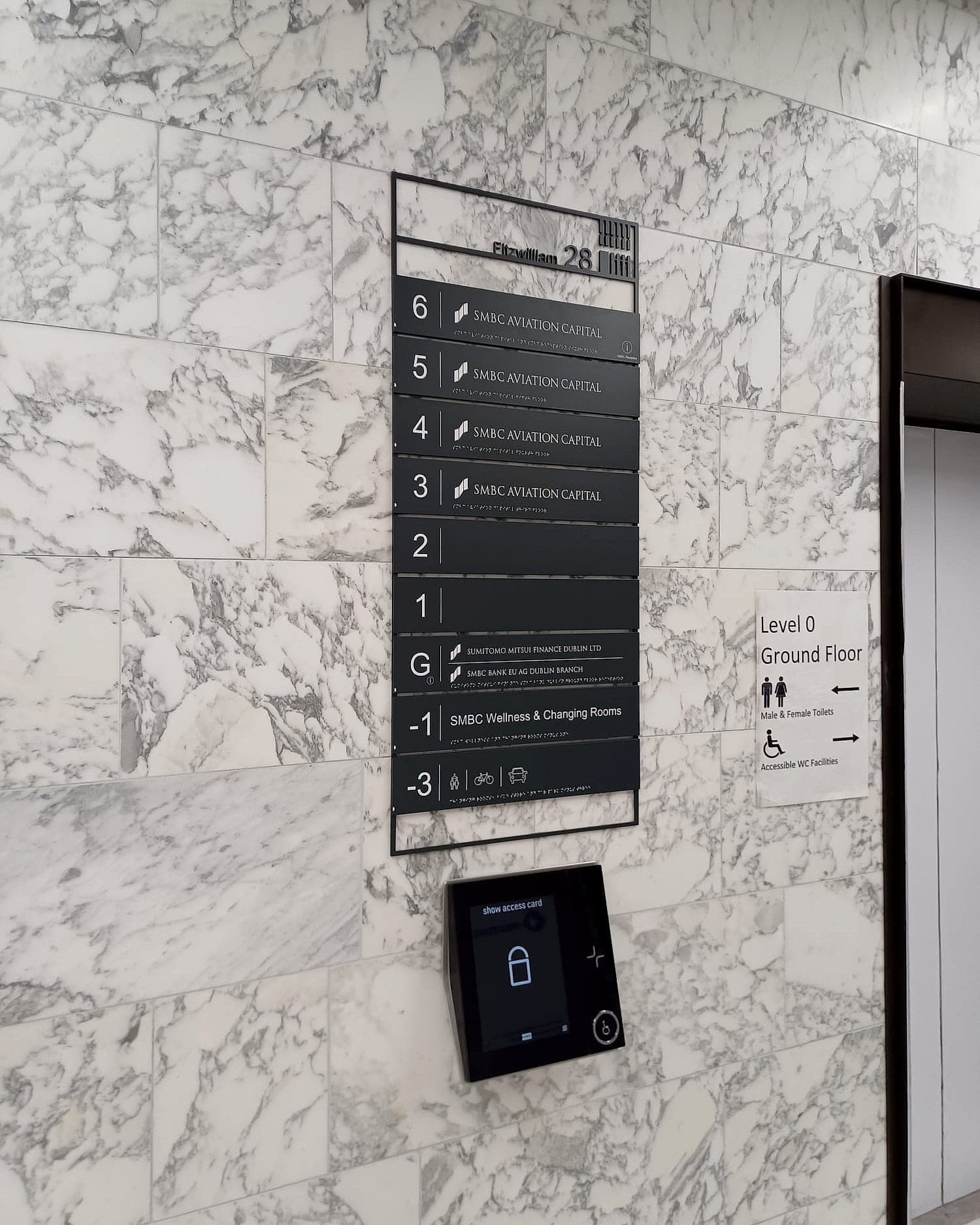 Level directory signage at SMBC in a lift lobby, listing levels minus three to six against their occupants, with tactile lettering and braille, mounted directly above a wall-mounted access-card reader