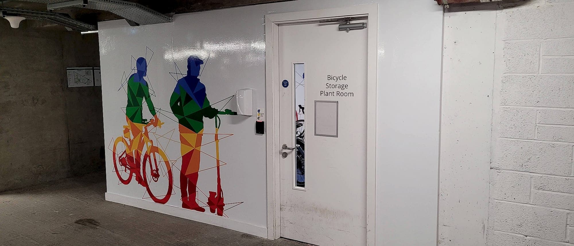 A white plant-room door in a basement services zone, with an access-control reader beside the handle and exposed cable tray and a sprinkler main overhead