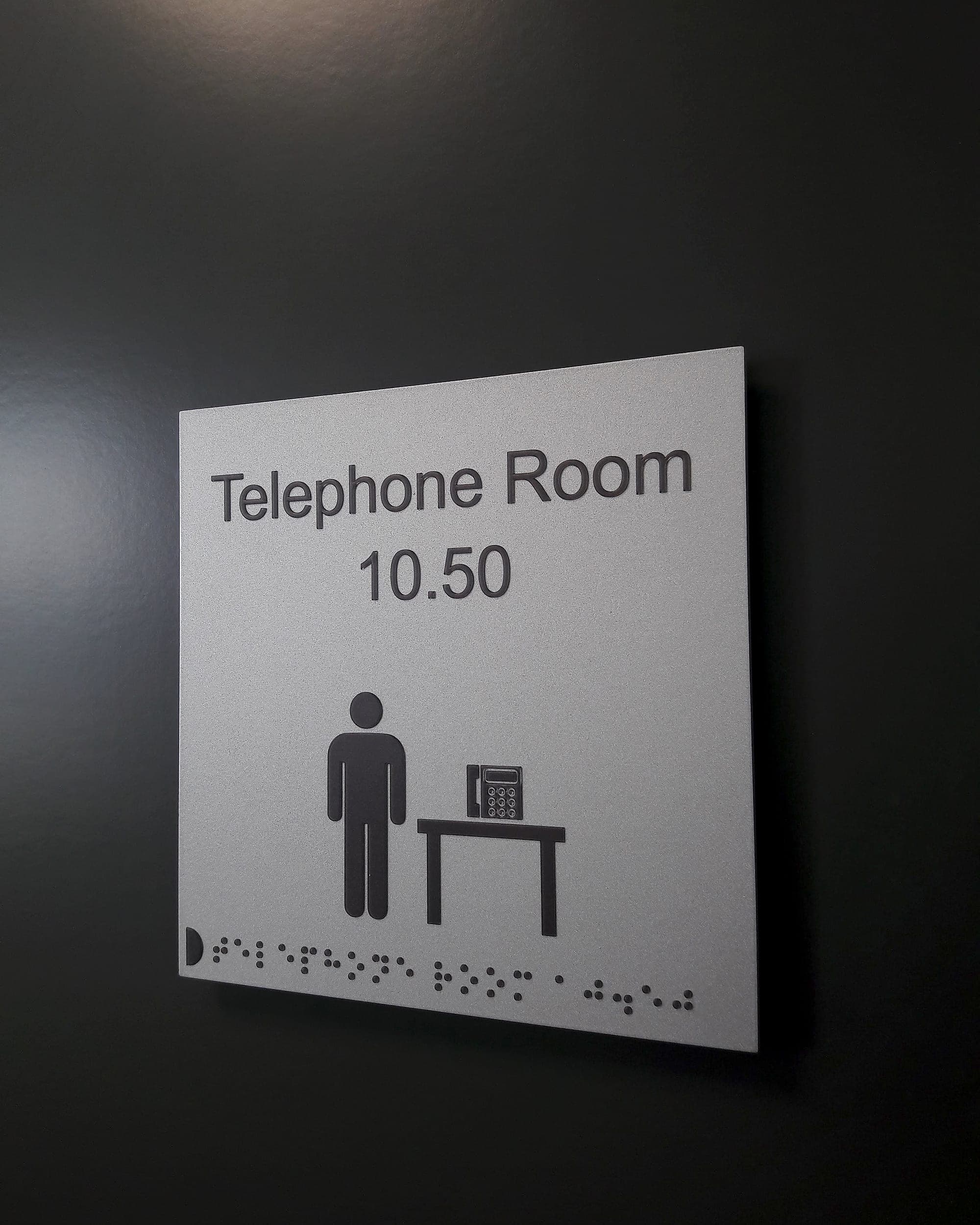 Room identification signage with the room name and number in raised tactile mixed-case lettering, a pictogram and a line of braille along the lower edge, on a light panel against a dark wall