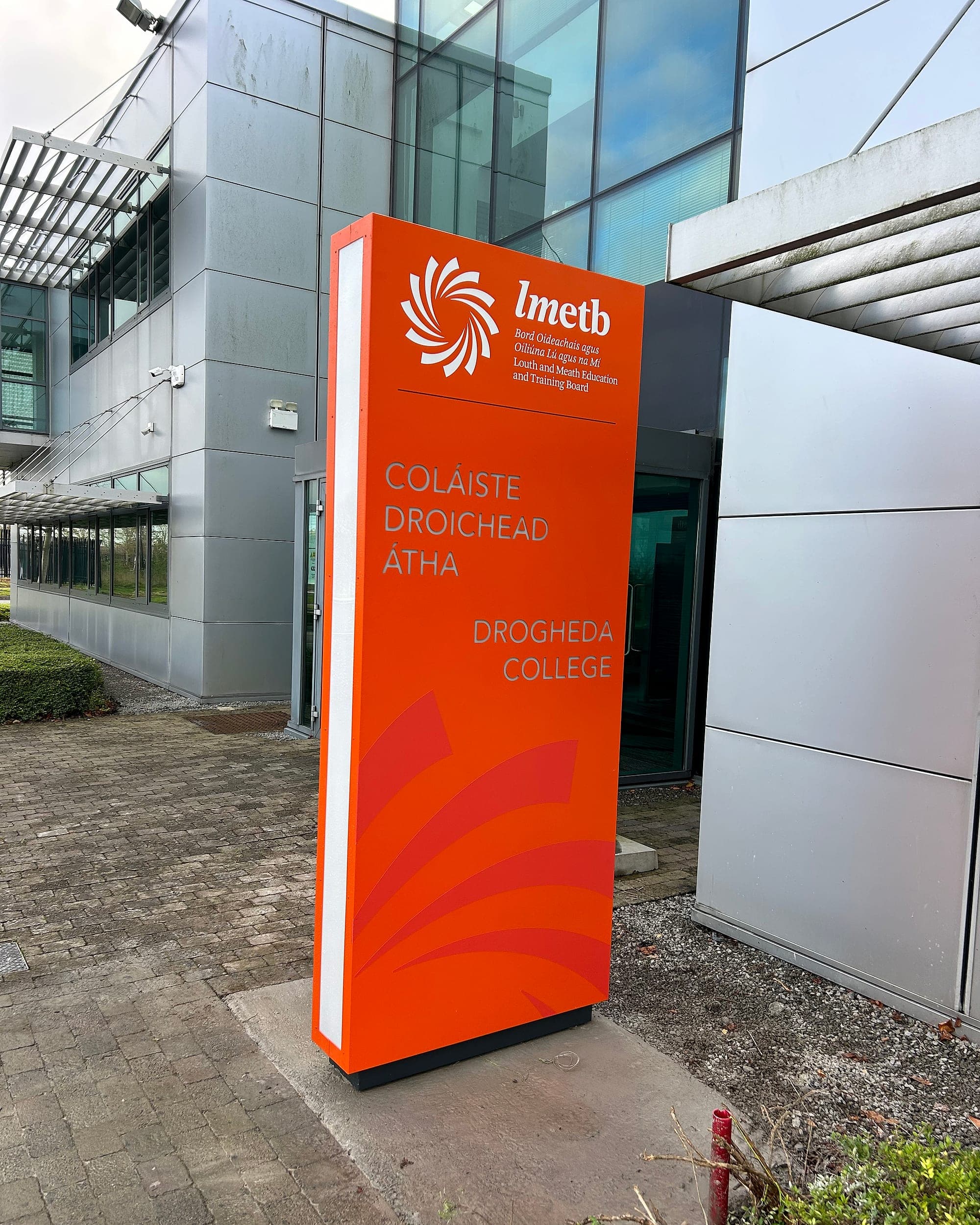 External entrance totem at Drogheda College: full-height orange panel carrying the bilingual Irish and English college name and the LMETB identity beside the main door