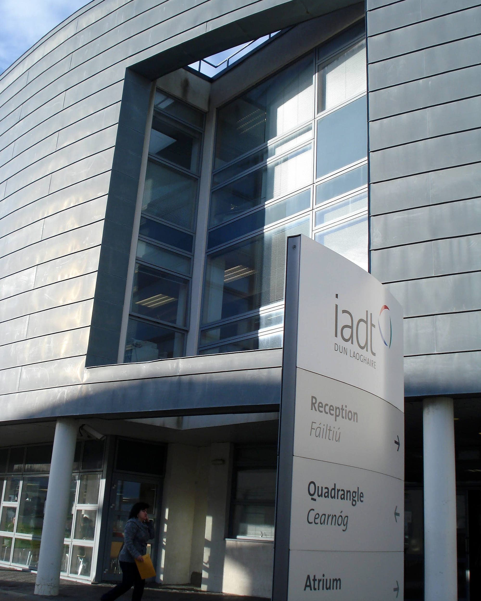 External directional totem outside a campus building at IADT Dún Laoghaire, listing Reception, Quadrangle and Atrium in English and Irish with arrows, set against a curved metal-clad façade