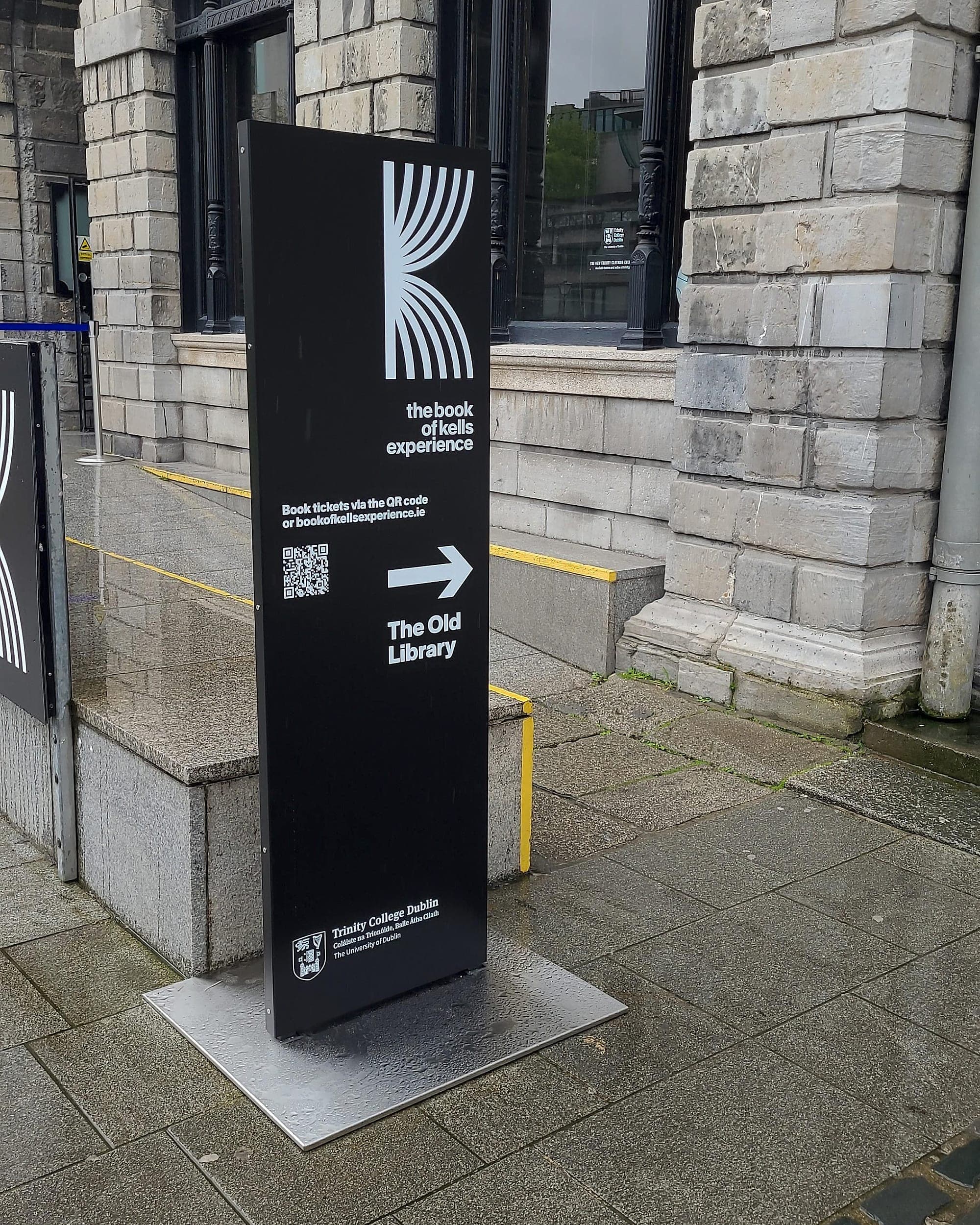 Signage maintenance and replaceability: black freestanding external totem directing visitors to the Book of Kells at the Old Library, Trinity College Dublin, standing on a stainless baseplate on wet granite paving
