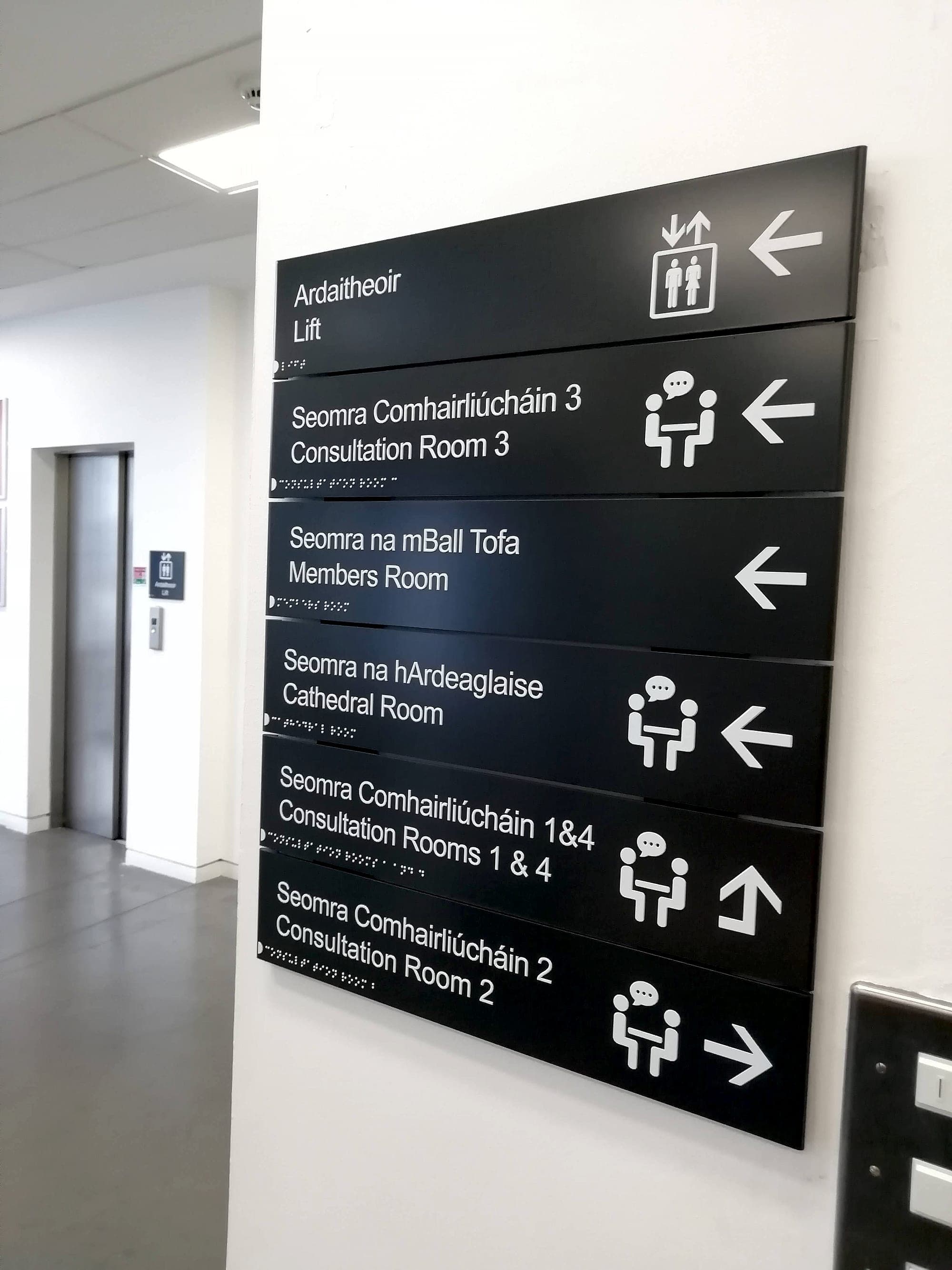 A lift lobby with high-contrast bilingual directional signage for visually impaired readers: white mixed-case Irish and English lettering, pictograms and arrows on charcoal panels, with a braille line beneath every destination, beside a lift