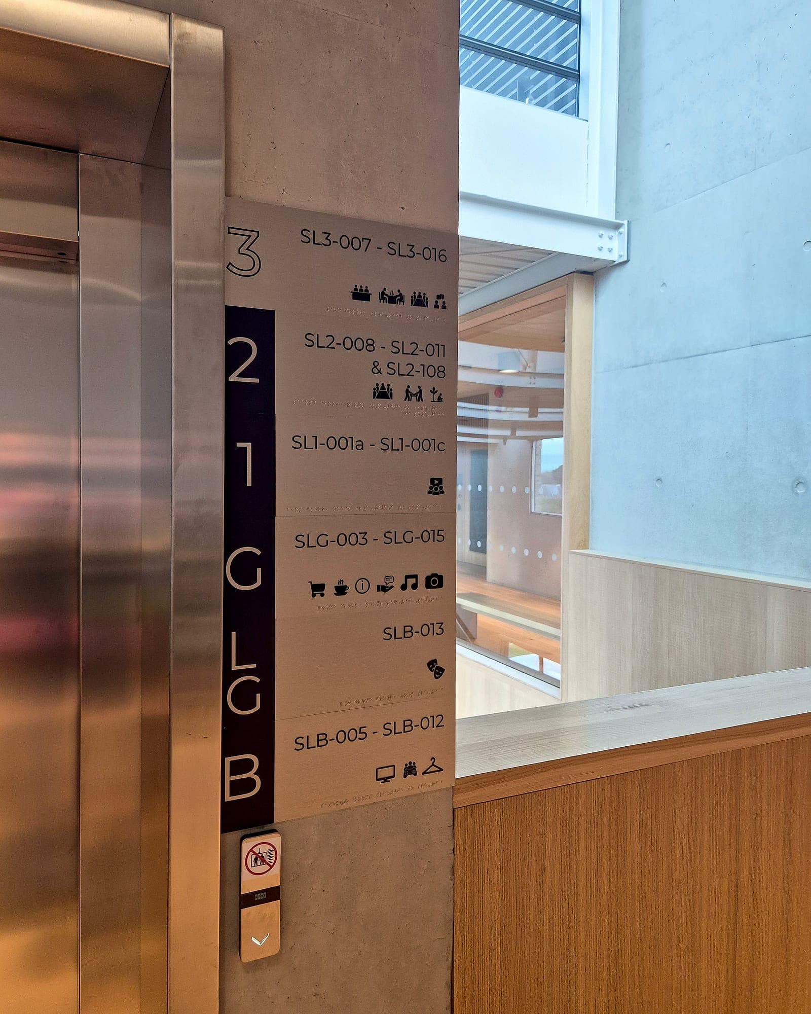 University of Limerick Student Union: level directory panel mounted on a concrete pier beside lift doors, listing room-number ranges by level with pictograms and braille, with a prohibition notice beside the lift call button