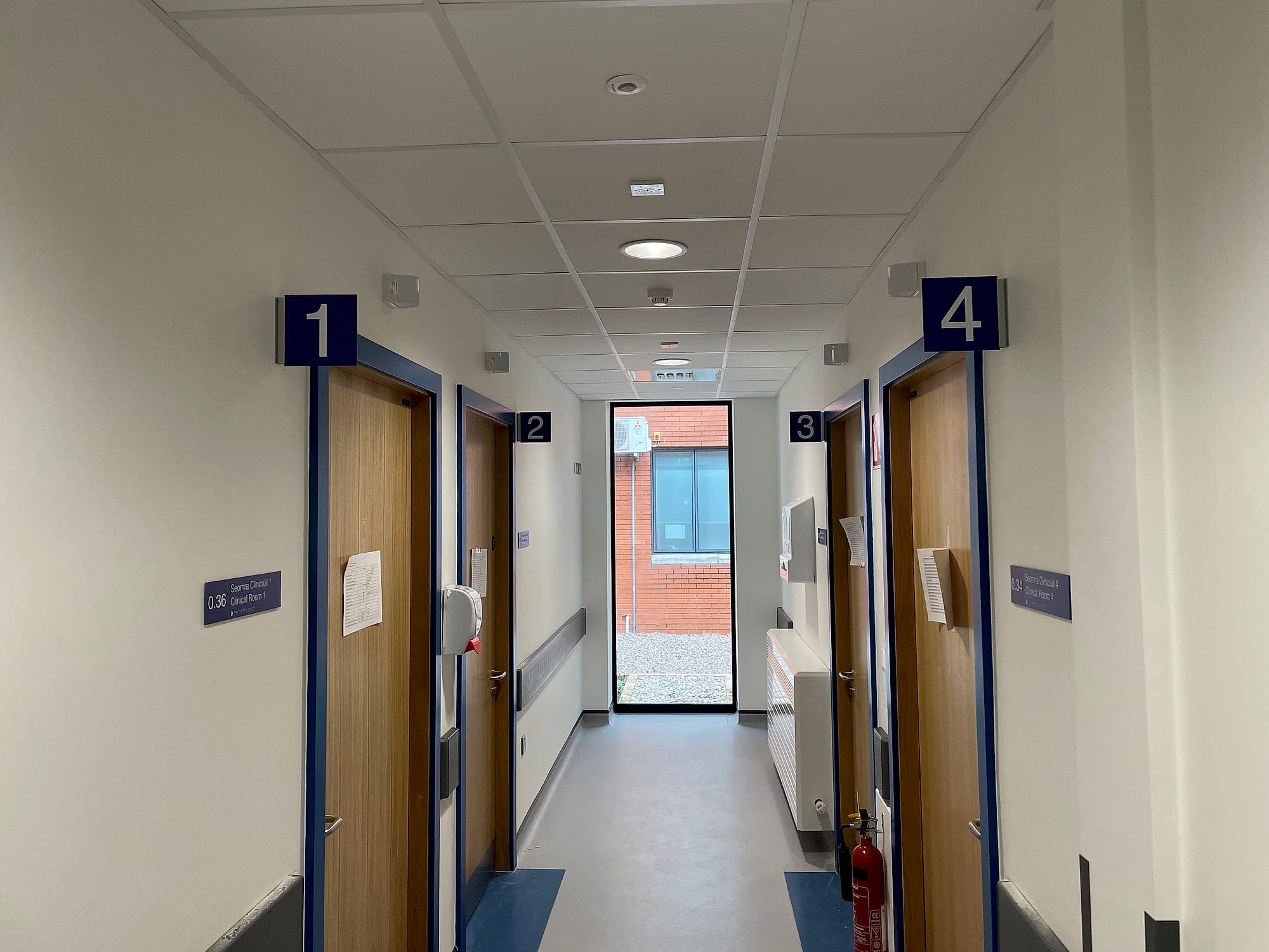 Medical centre signage in a clinic corridor at South Infirmary Victoria University Hospital: projecting numerals 1 to 4 over consulting-room doors with bilingual Irish and English door signs carrying room numbers and braille
