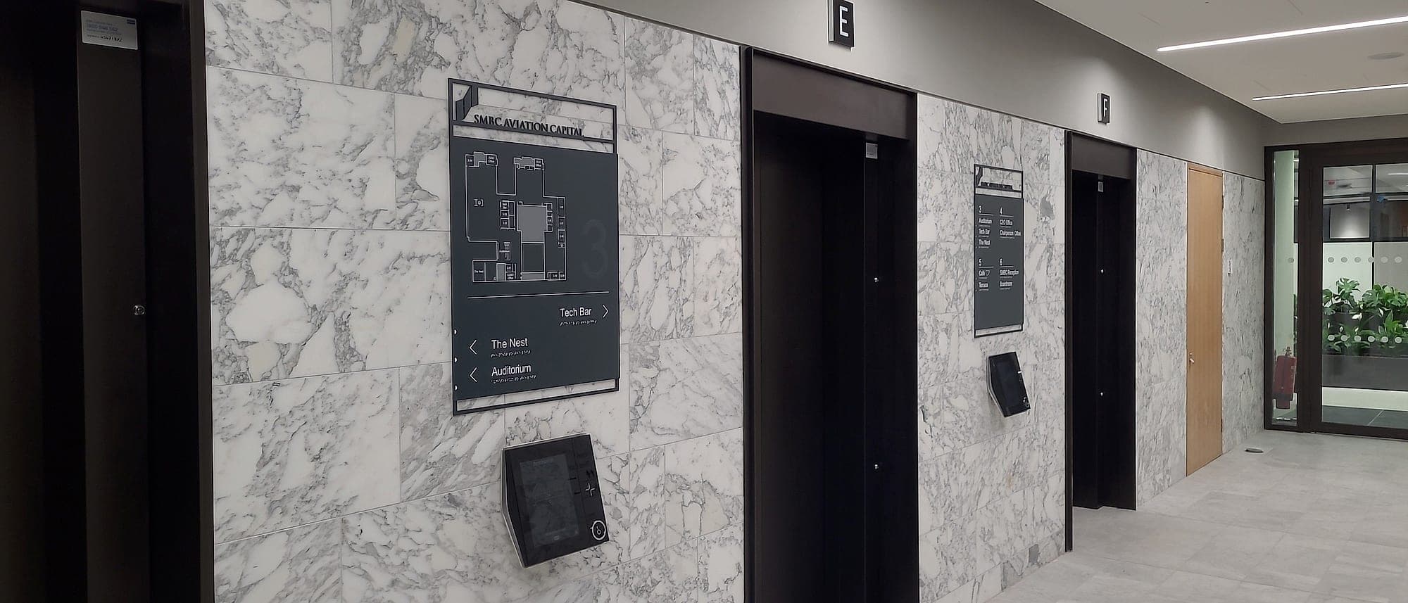 SMBC: office lift lobby with a layered floor-plan and directional sign set into a marble wall, tactile and braille lettering, a wall-mounted screen beside it, and lift-core identification above the doors
