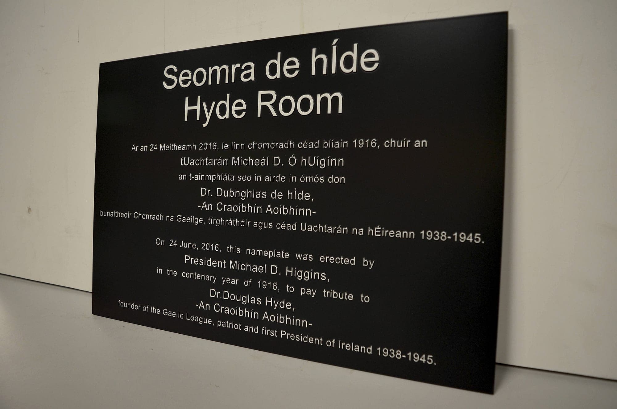 Bilingual Irish and English room nameplate with raised, painted lettering on a dark panel, photographed against a plain wall