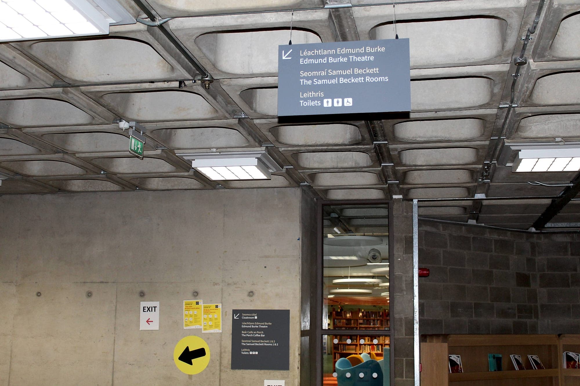 Safety signage and wayfinding sharing one wall: a suspended bilingual Irish and English directional sign, a green running-man escape luminaire on the same ceiling, and taped paper exit notices and a stick-on arrow below