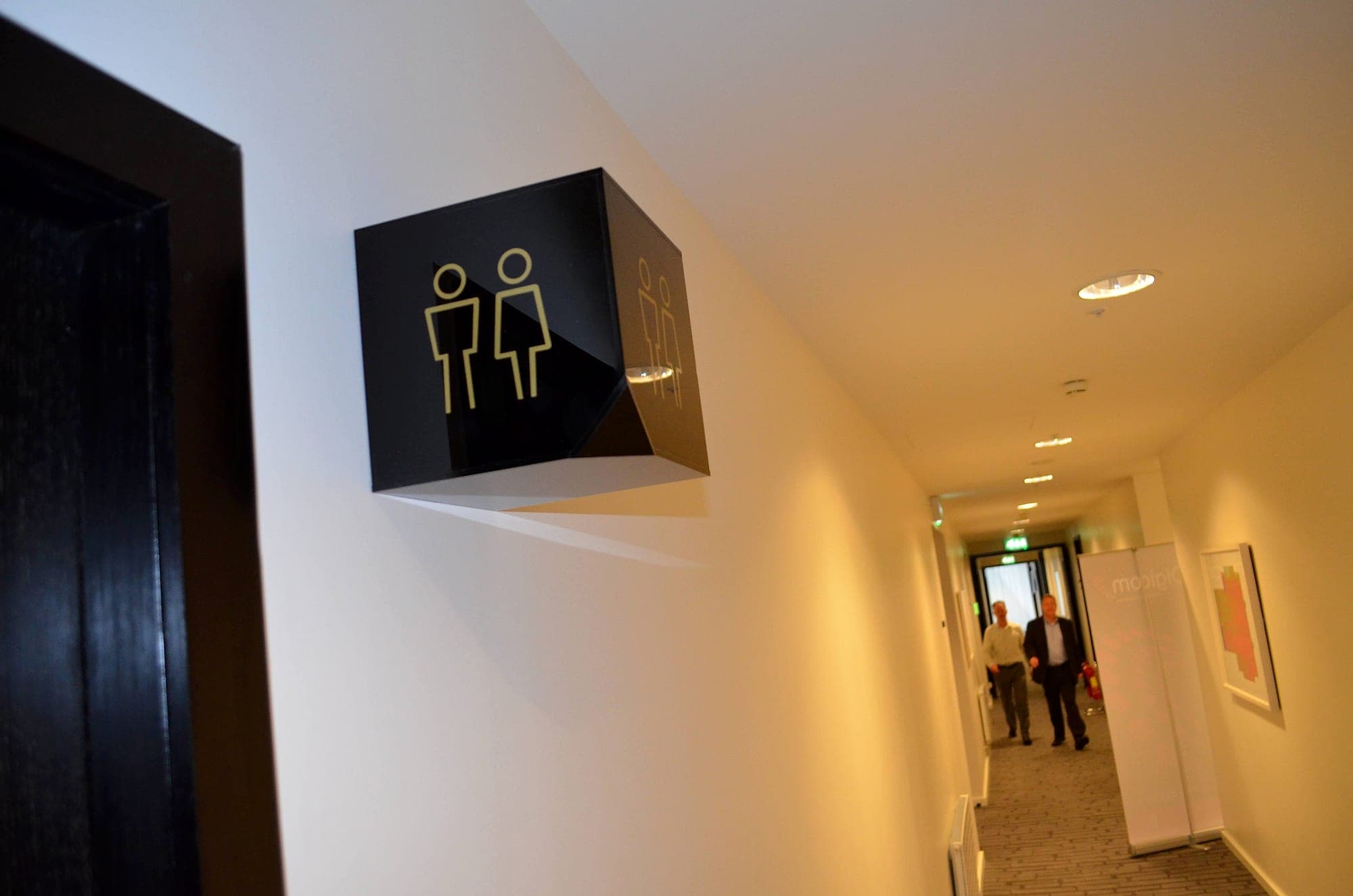 Projecting cube toilet sign with gold pictograms in a hotel corridor at The Marker Hotel, seen from below along the line of approach, showing its projection from the wall and the headroom beneath it