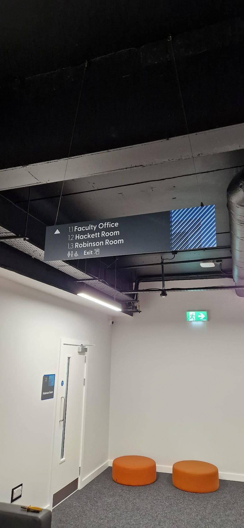 Suspended directional wayfinding sign at a corridor junction in CCT College Dublin, listing three numbered destinations (1.1 Faculty Office, 1.2 Hackett Room, 1.3 Robinson Room) with an arrow, toilet and accessibility pictograms, and the matching numbered door sign on the room below