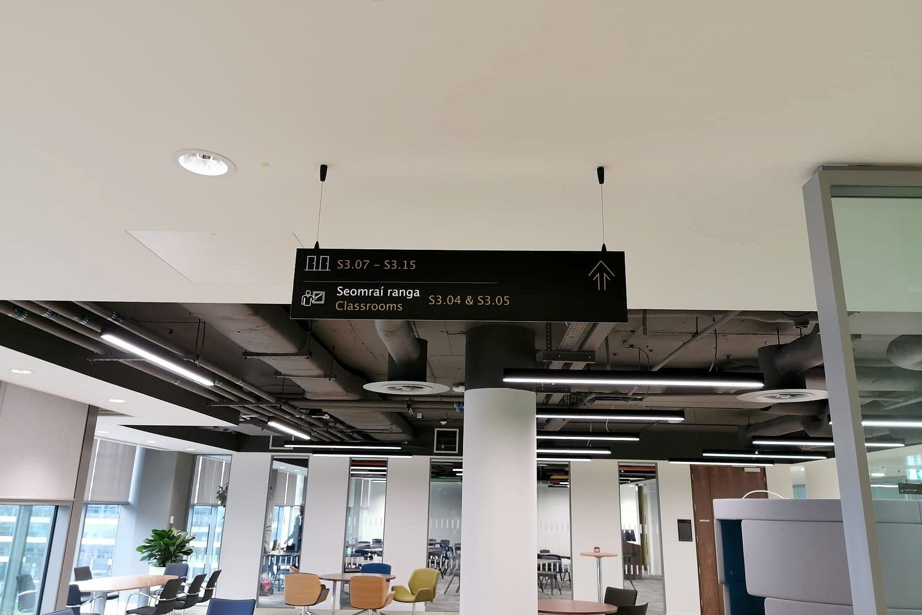 Suspended bilingual Irish and English directional sign on an open teaching floor, listing room codes S3.07 – S3.15 and Seomraí ranga / Classrooms S3.04 & S3.05 with an upward arrow