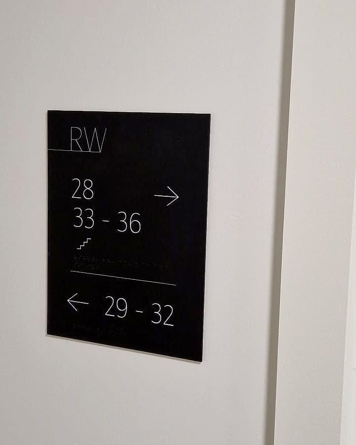 Multi-sensory wayfinding at Rathborne Wharf, Dublin: black corridor directional sign with white flat numbers, a stair pictogram and a braille line beneath each destination