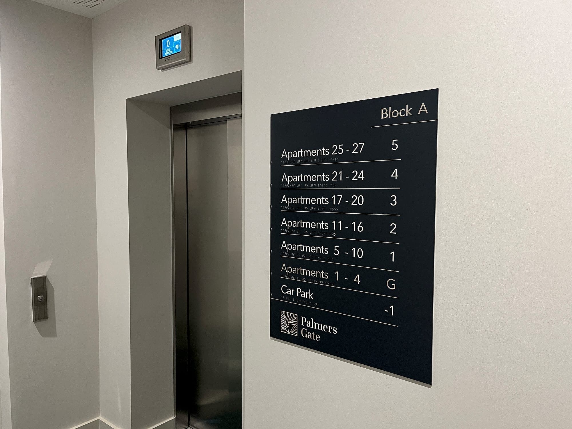 Palmers Gate. Certified signage at a residential lift lobby: tactile level directory with braille beneath every line, listing apartment ranges by floor beside the lift doors
