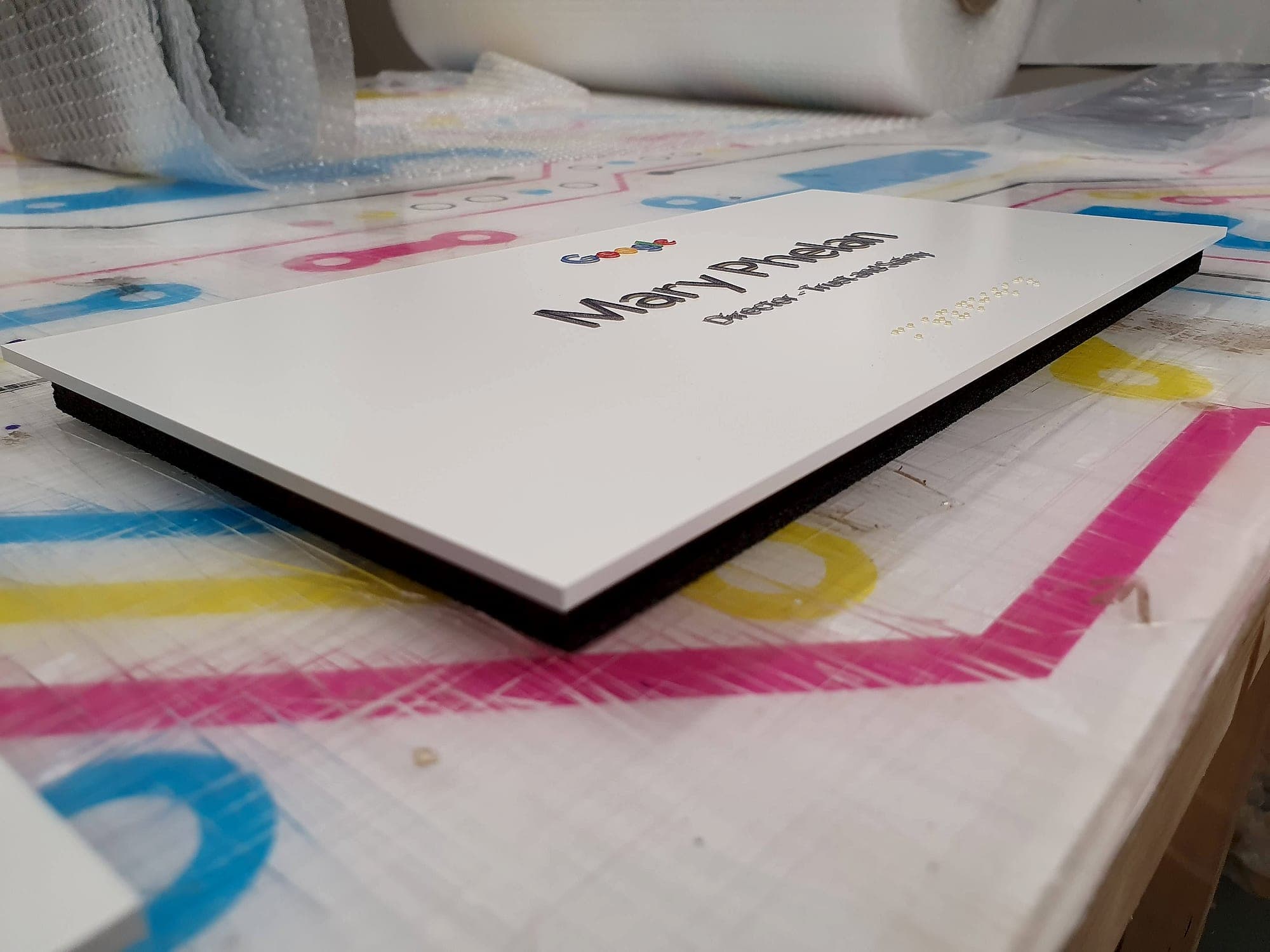 Sign manufacturing on the workshop bench: a finished white office door sign with raised tactile lettering and braille, its dark panel core visible along the cut edge, resting on an ink-marked production table