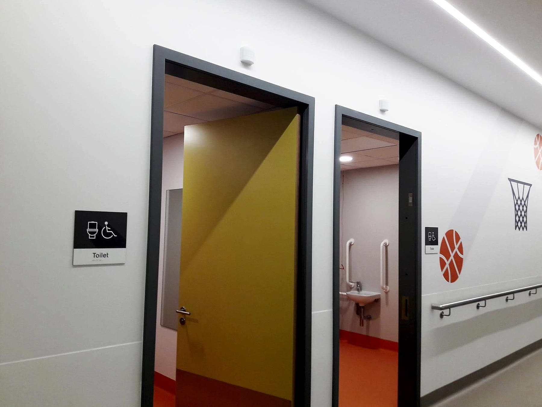 Accessible toilet signage in a hospital corridor at the National Rehabilitation Hospital: high-contrast pictogram panels beside sports-themed wall graphics and a handrail