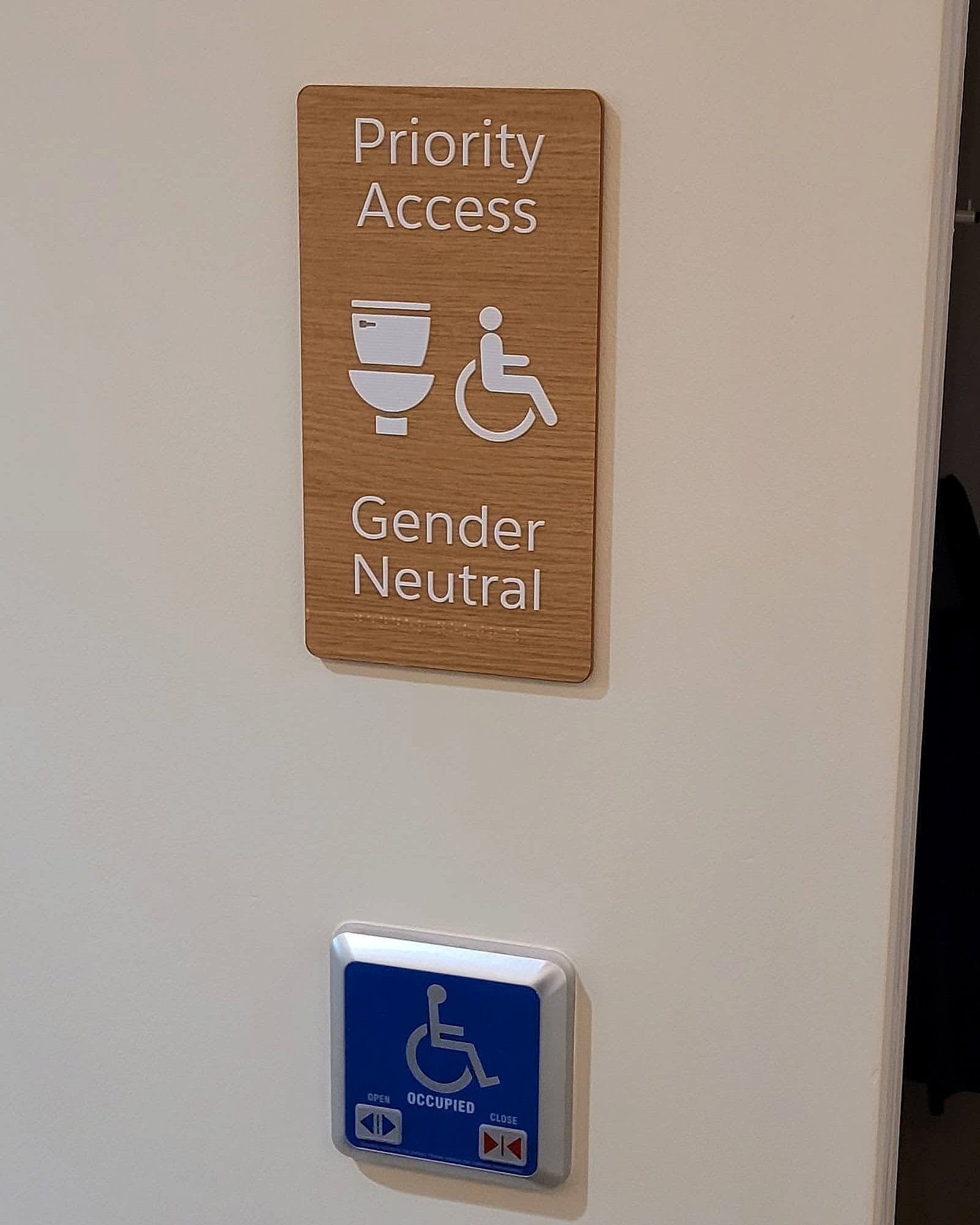 Salesforce: accessible toilet signage on an office wall, with a timber-faced priority access and gender neutral sign with pictograms, raised lettering and braille, above a blue accessible door control