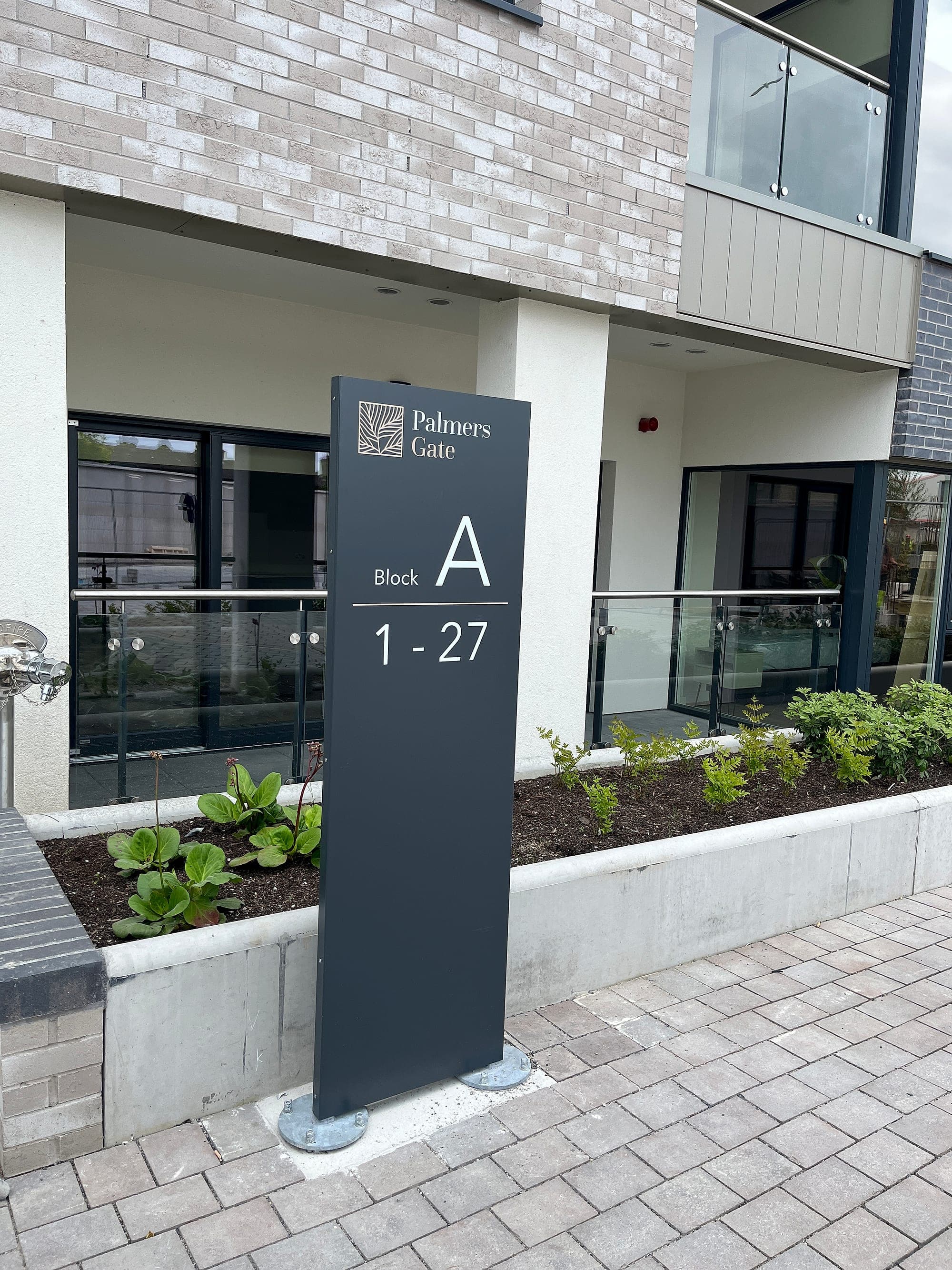 Dark grey aluminium totem at the entrance to Block A, Palmers Gate, showing the scheme logo and apartments 1 to 27, set in a planted bed in front of the apartment building.