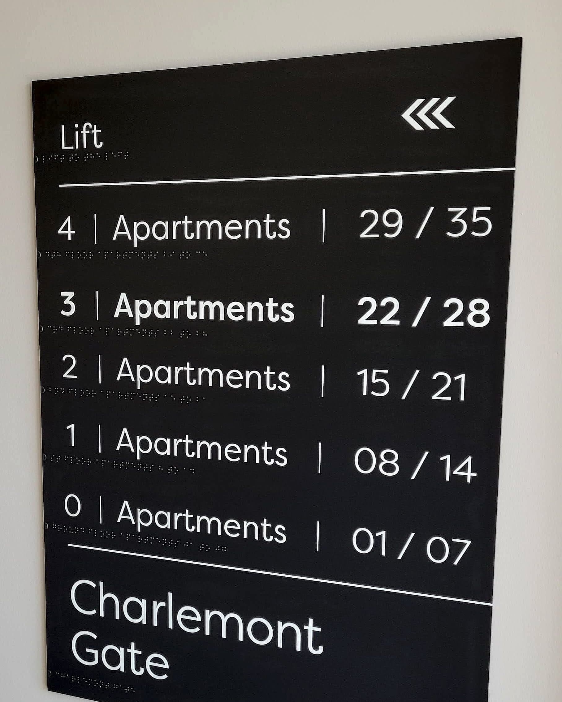 Apartment block floor directory at Charlemont Gate: dark lift-lobby board listing floors four down to zero against apartment number ranges, with the current floor picked out in bold and braille under every line