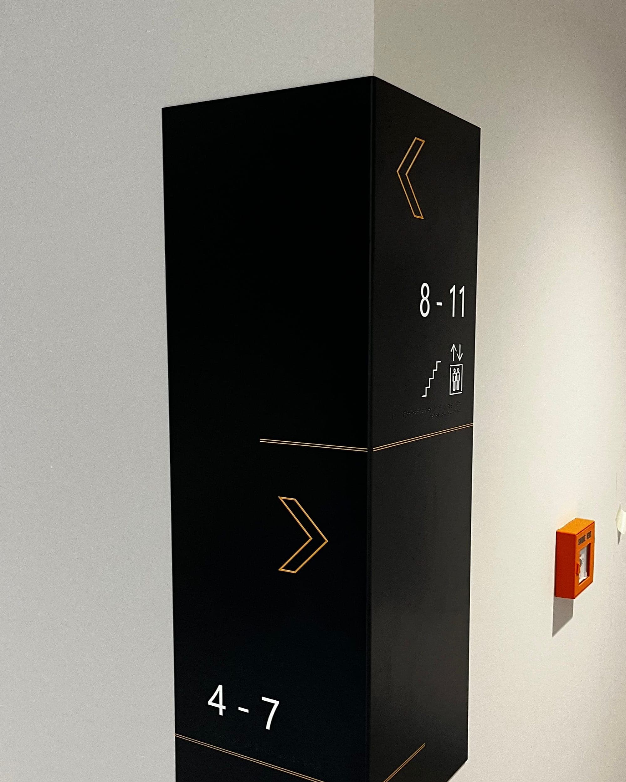Millers Glen: apartment stair core flat number signage, a black double-sided projecting blade sign splitting flats 8-11 one way and 4-7 the other, with stair and lift pictograms, beside a break-glass call point