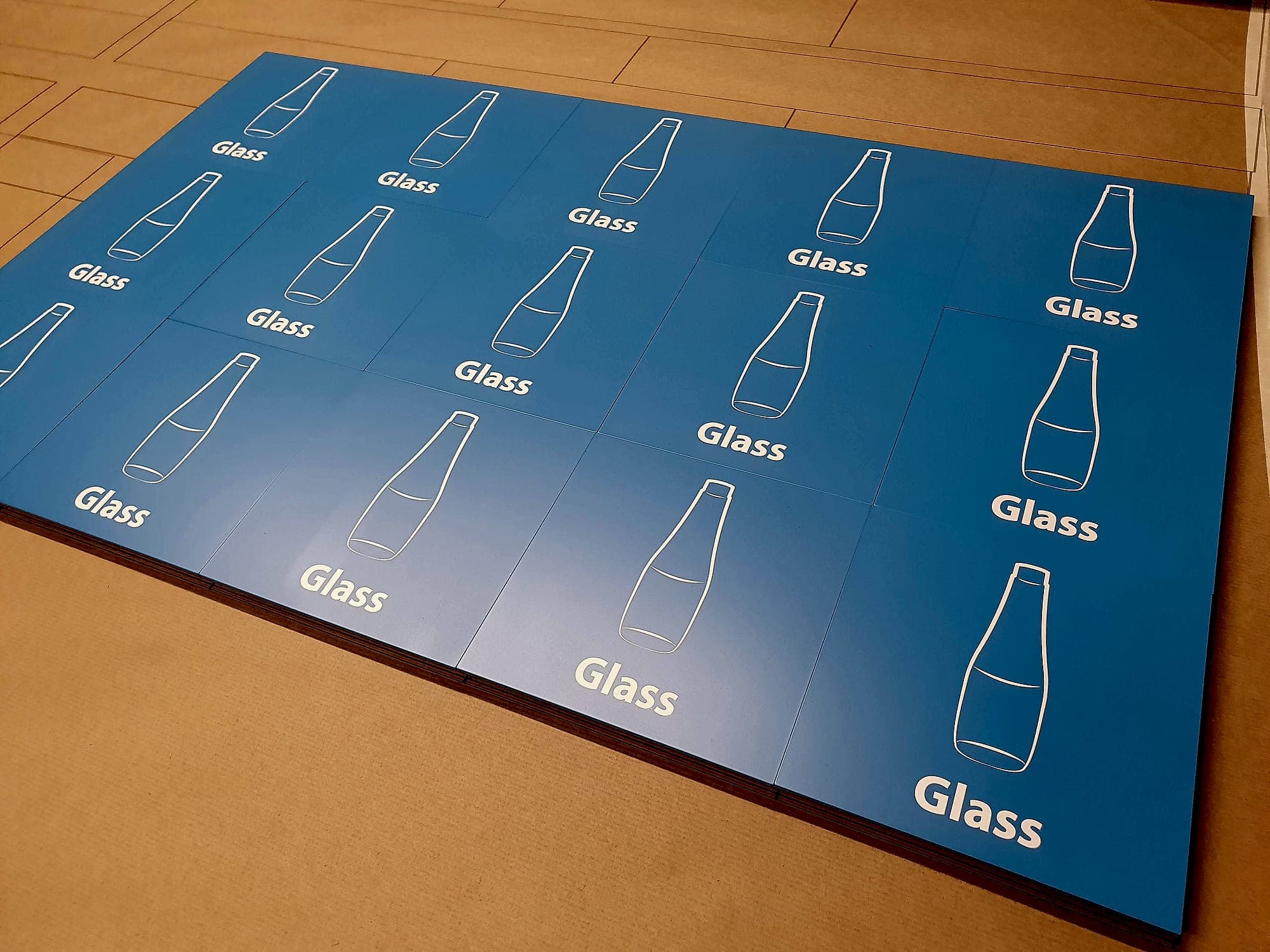 Batch sign manufacturing for Apple at Hollyhill, Cork: a stack of identical printed blue recycling identification panels laid out on the workshop floor, each with a glass-bottle pictogram and the word Glass