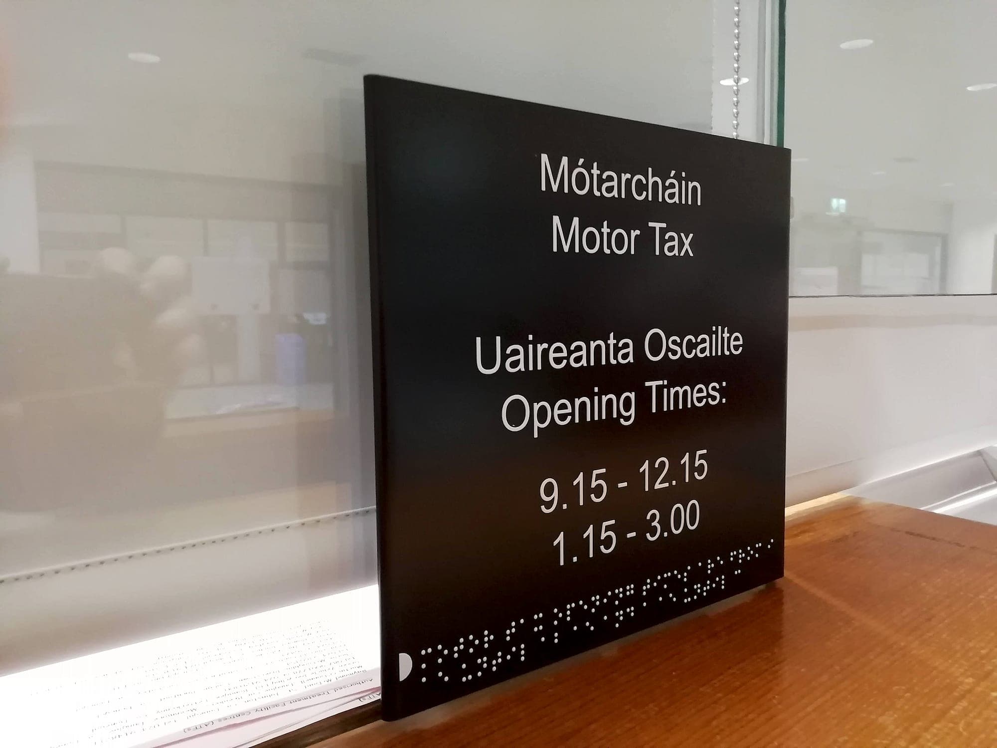 Motor Tax Office: bilingual Irish and English motor tax counter sign in white lettering on a matt black panel with braille along the lower edge, standing at a public service counter