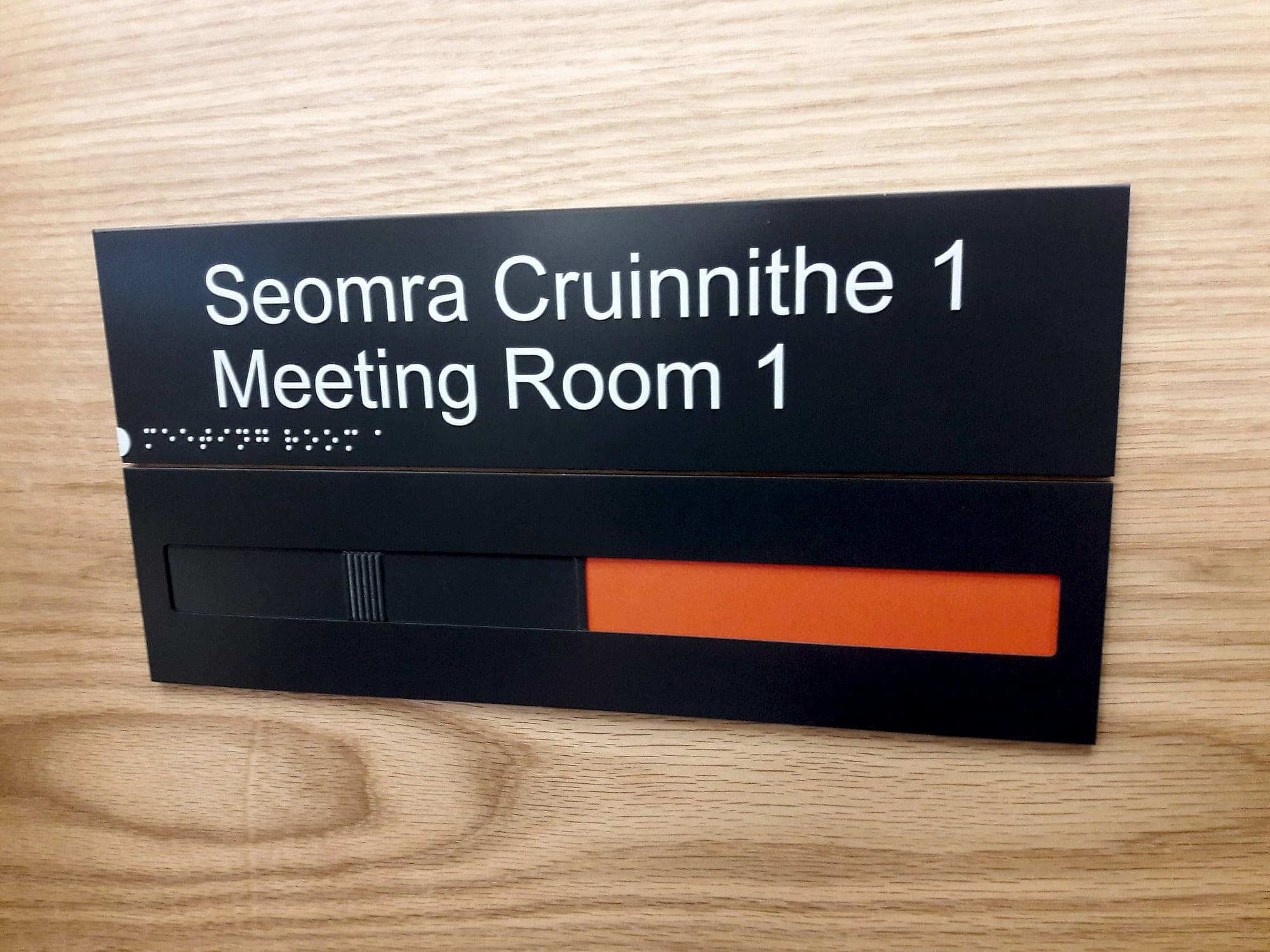 Bilingual Irish and English meeting room door sign reading Seomra Cruinnithe 1 / Meeting Room 1 in white on matt black, with braille along the lower edge and a sliding vacant and engaged indicator below