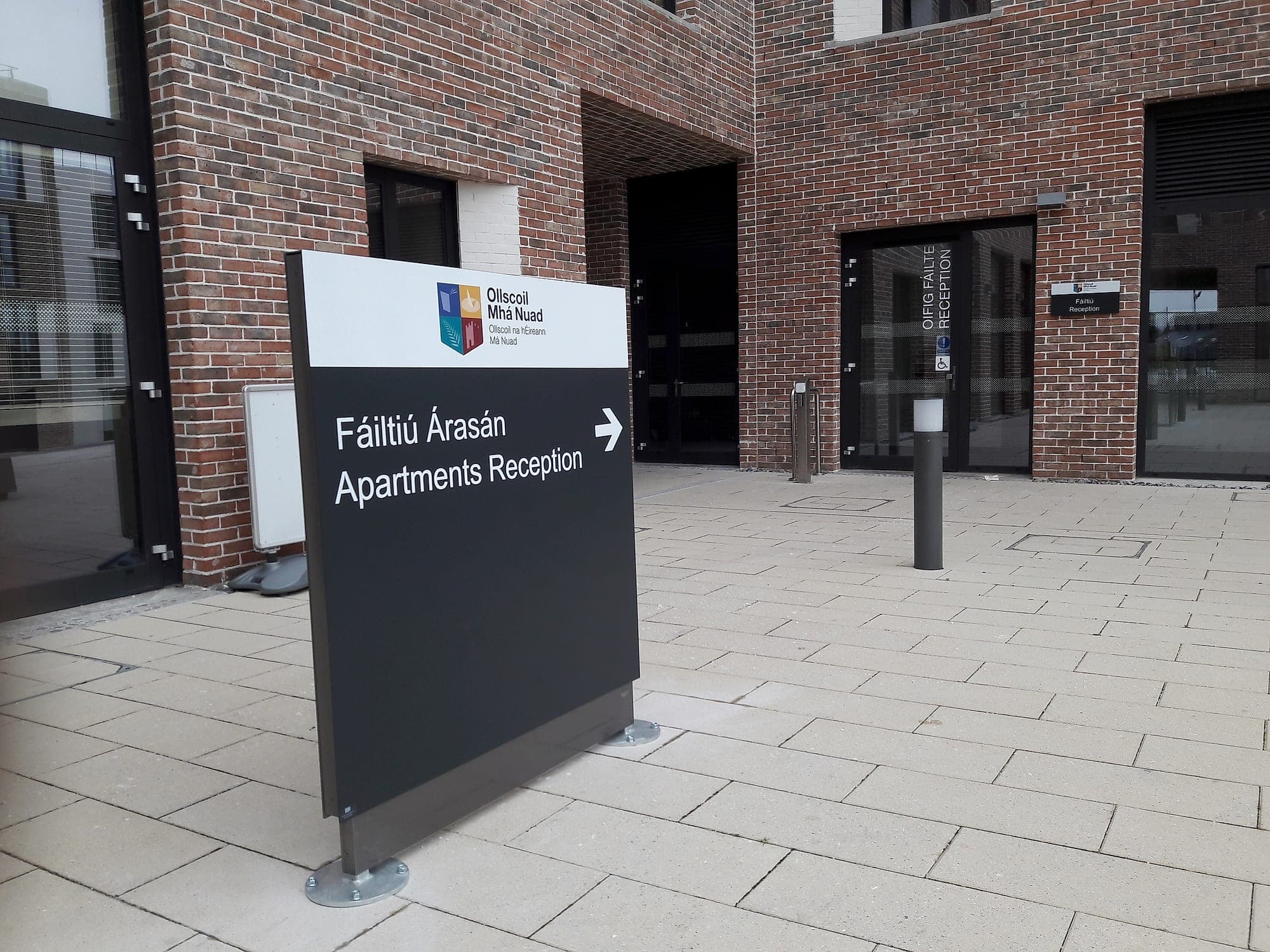 Signage compliance and brand colour on a Maynooth University campus totem: the full-colour crest on a white header band above a bilingual Irish and English directional panel in white on near-black