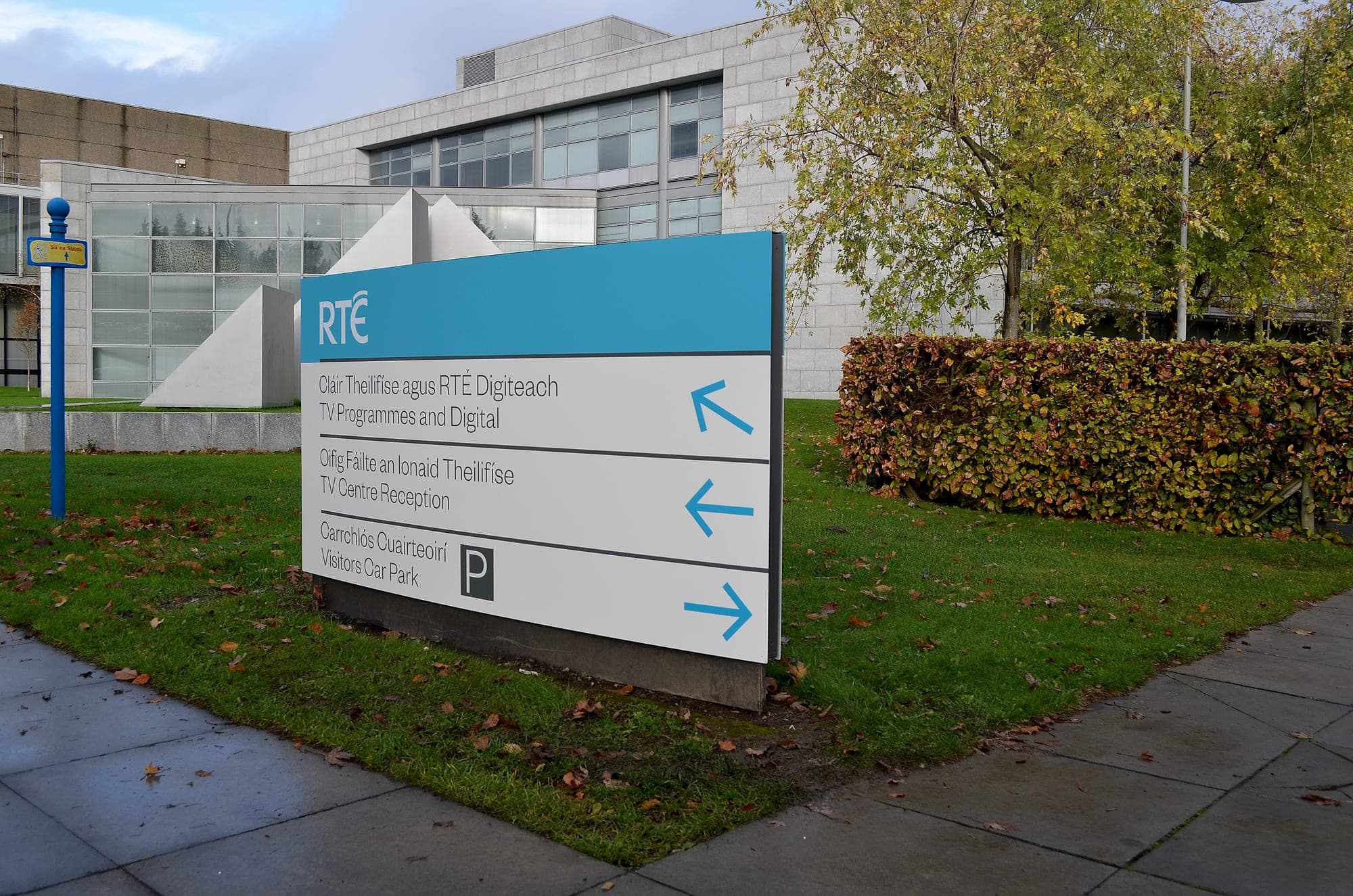 Bilingual external directional sign on the RTÉ campus, the RTÉ mark on a brand-blue header above a white panel listing TV Programmes and Digital, TV Centre Reception and Visitors Car Park in Irish and English with blue arrows