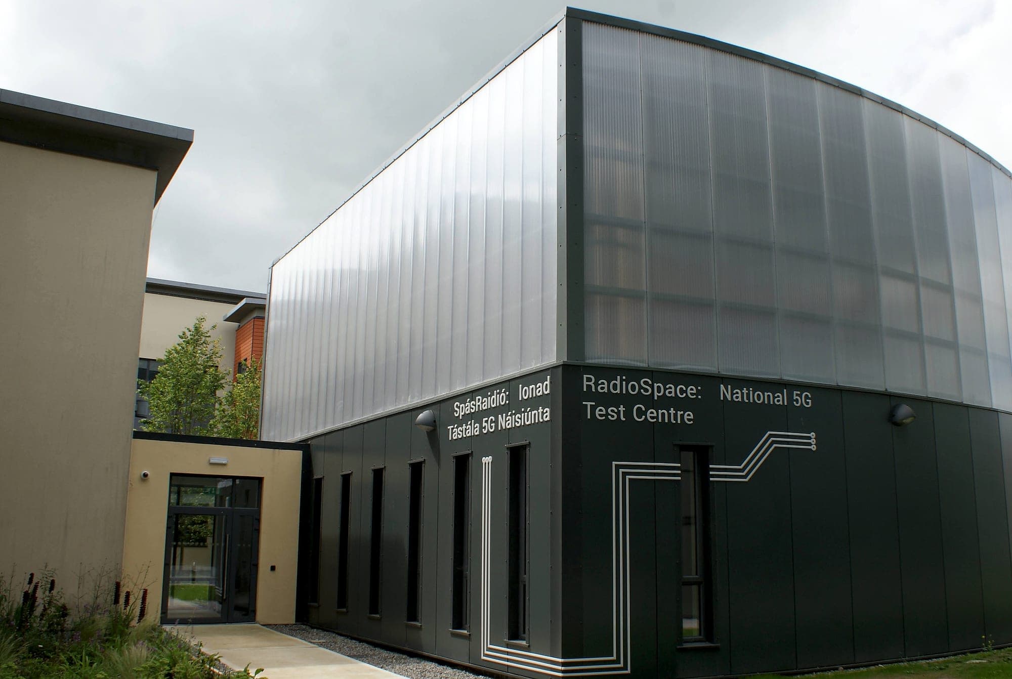 The National 5G Test Centre, with bilingual building naming lettered into a dark-clad research building. RadioSpace: National 5G Test Centre in English across one elevation and SpásRaidió: Ionad Tástála 5G Náisiúnta in Irish on the return