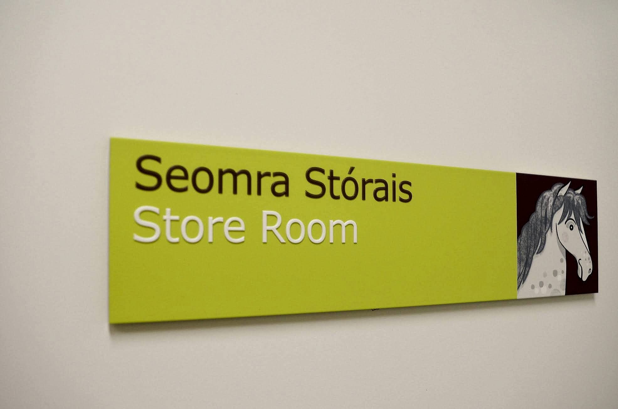 Bilingual room signage at Temple Street Children's Hospital, Dublin: a lime panel reading Seomra Stórais above Store Room in raised lettering, with a horse illustration in a plum block at the right end