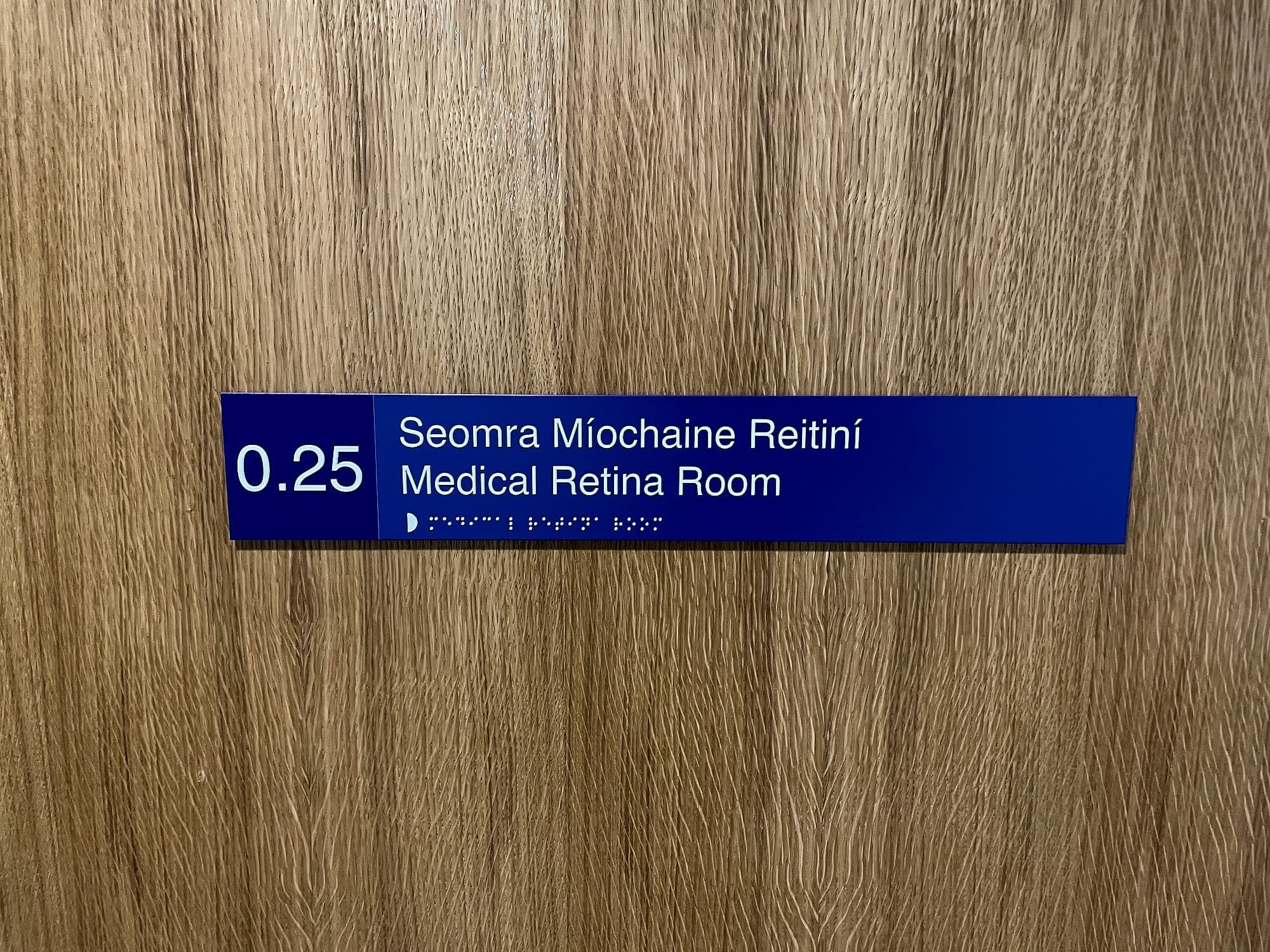 Bilingual clinic room door sign at South Infirmary Victoria University Hospital: room number 0.25 with the room name in Irish and English and braille beneath, mounted on a timber door