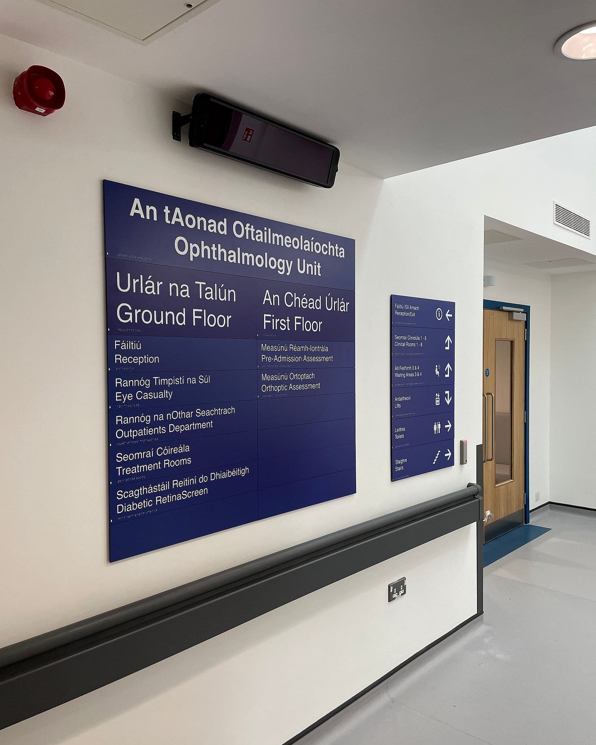 Bilingual Irish and English department directory at South Infirmary Victoria University Hospital listing an ophthalmology unit's rooms by ground and first floor, with a separate directional panel beside it for reception, clinical rooms, waiting areas, lifts, toilets and stairs