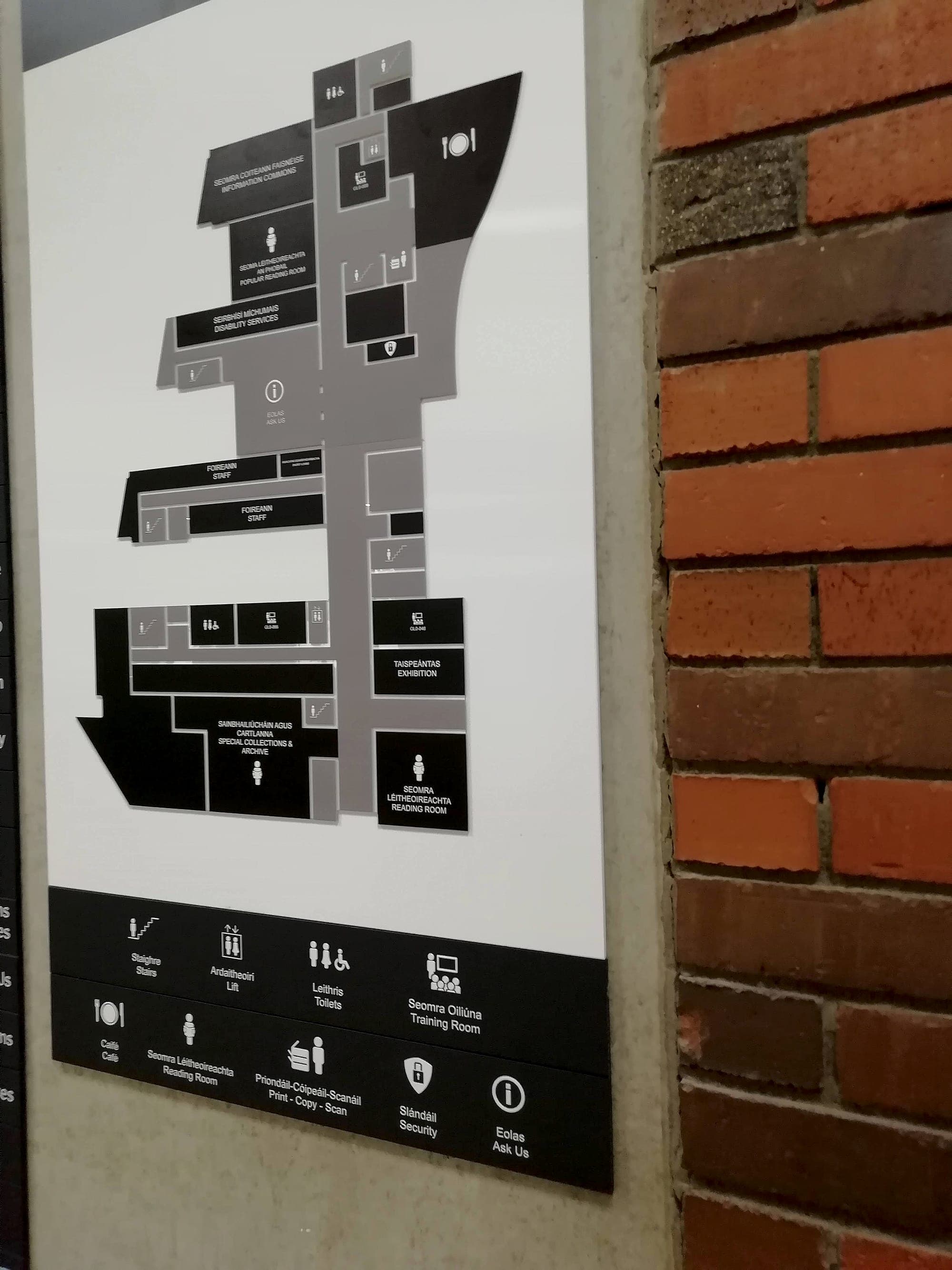 Bilingual Irish and English you-are-here floor plan sign in the Glucksman Library, with rooms picked out in black, room codes such as GLD-035 printed on the plan, and a pictogram key beneath