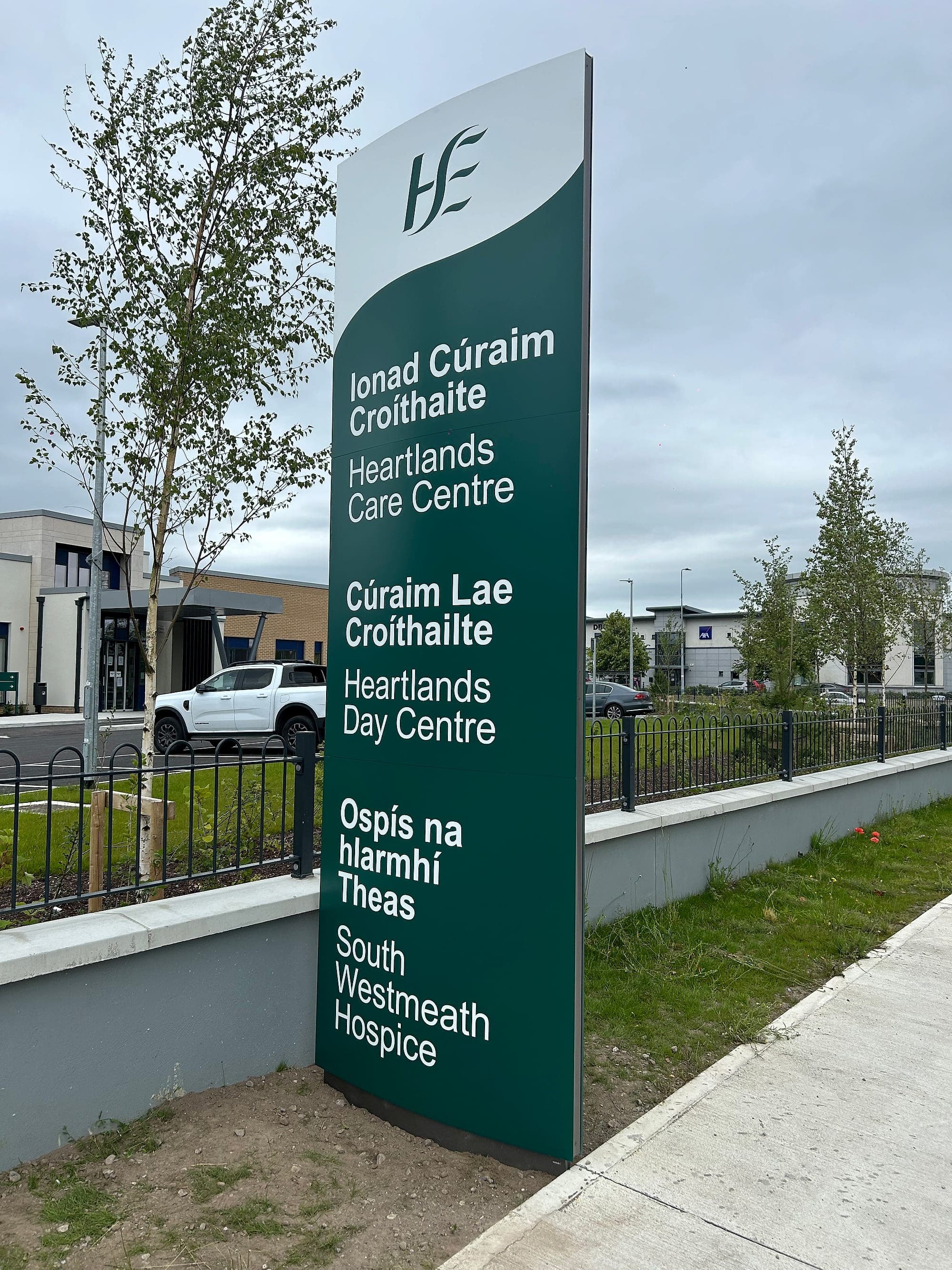 Bilingual Irish and English entrance totem naming a care centre, day centre and hospice: a folded and painted monolith set on a concrete foundation at a healthcare site entrance