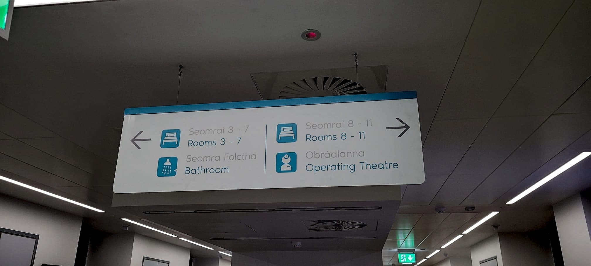 Bilingual Irish and English directional hospital signage suspended at a corridor junction in the Rotunda Hospital, Dublin, with pictograms for rooms, bathroom and operating theatre