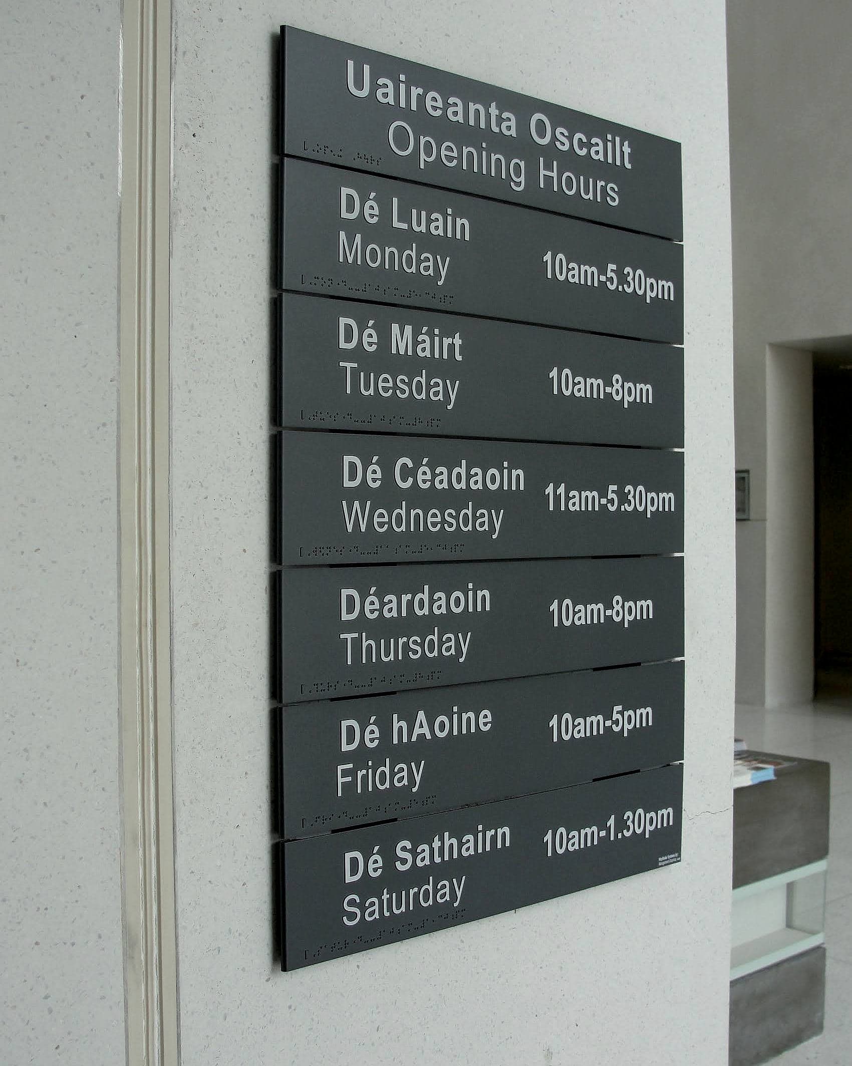 Opening hours as a wayfinding sign: bilingual Irish and English opening hours sign in a public library lobby, with Irish above English on every line and braille strips below each row