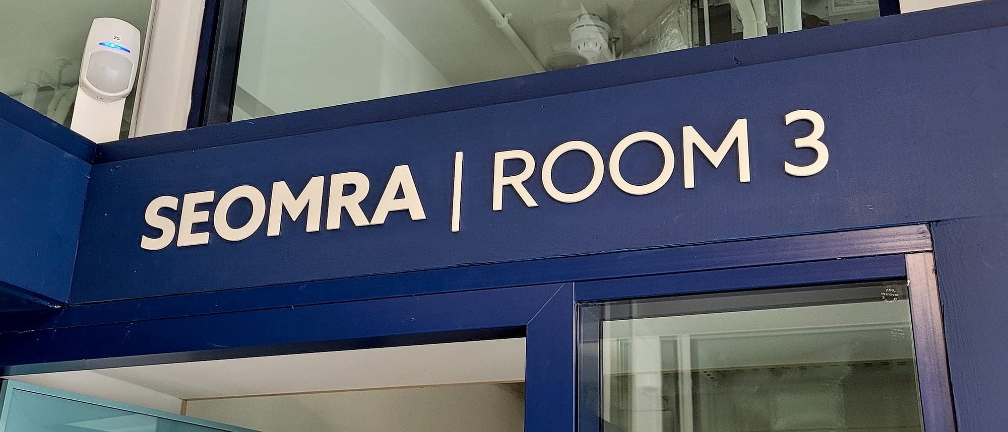 Bilingual Irish and English room identification lettering reading SEOMRA | ROOM 3 at the Garda station on O'Connell Street, individually applied in cream on a deep blue door head