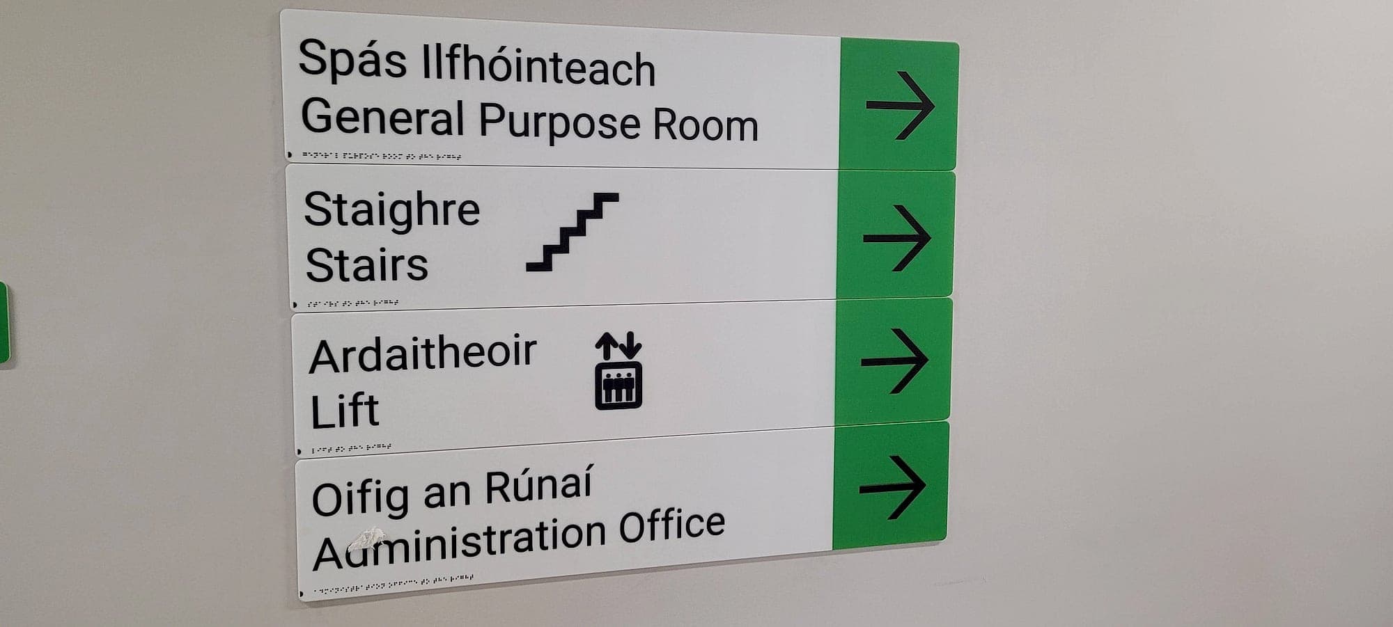Bilingual school wayfinding signage: a stack of four Irish and English directional panels with arrows and braille for General Purpose Room, Stairs, Lift and Administration Office