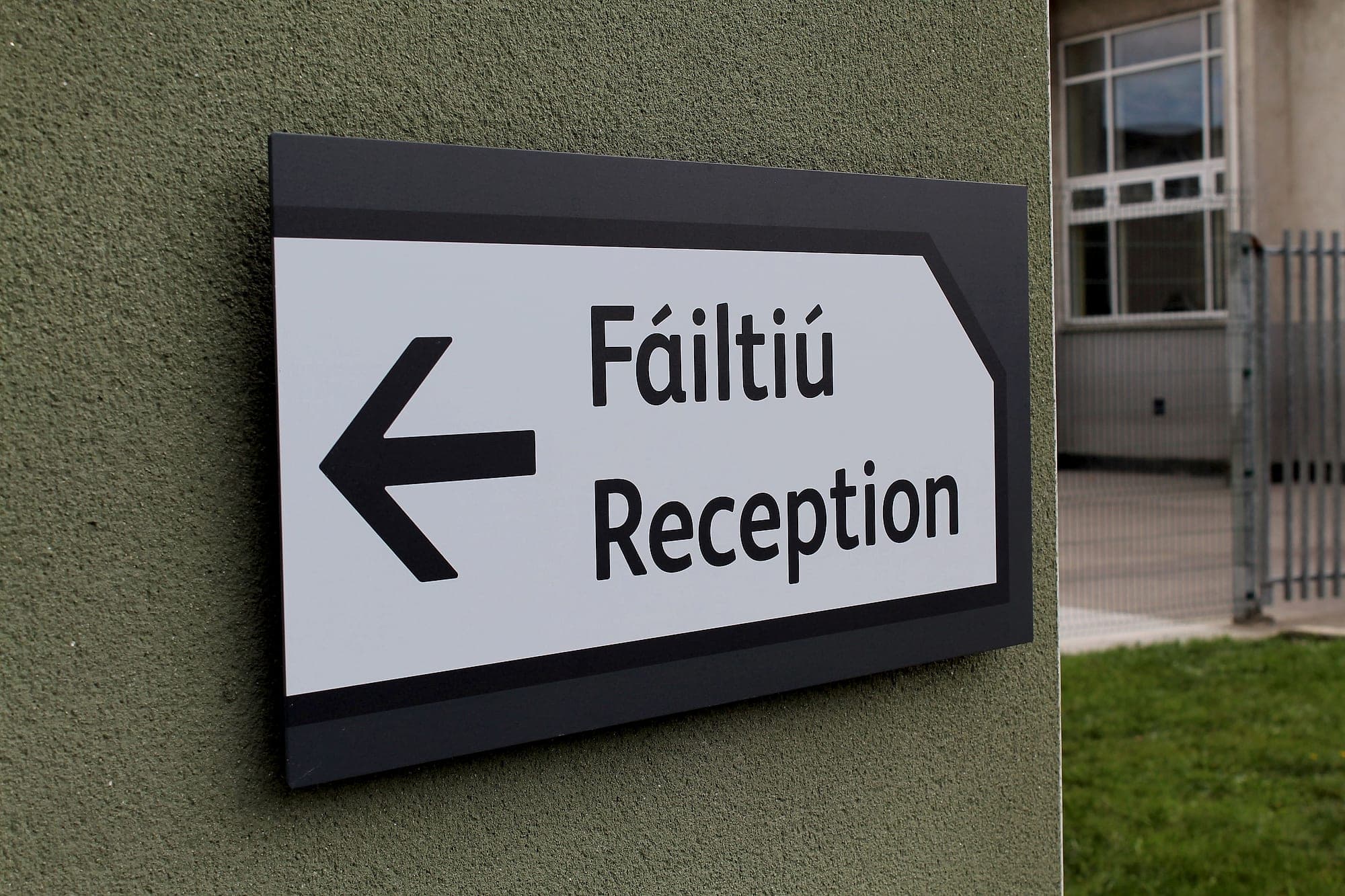Bilingual Irish and English school signage at Scoil Assaim: an external directional wall sign reading Fáiltiú / Reception with a left arrow, mounted beside the school's pedestrian gate